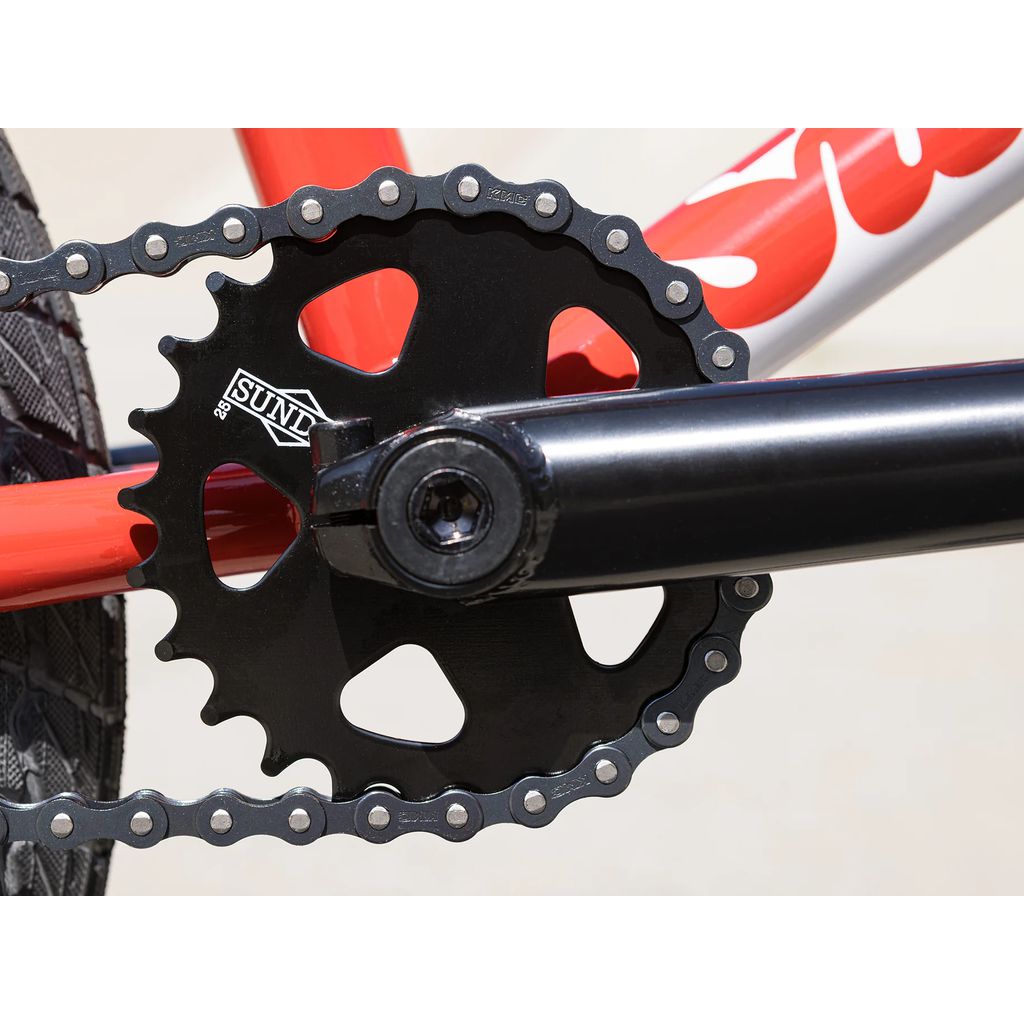 Close-up of a black crankset with a chain on a red bicycle frame. The crankset, emblazoned with the brand logo "SUNY," is part of the 2025 Sunday Primer 18 Inch Bike featuring Pro-Style geometry and Odyssey Springfield brakes for optimal performance.