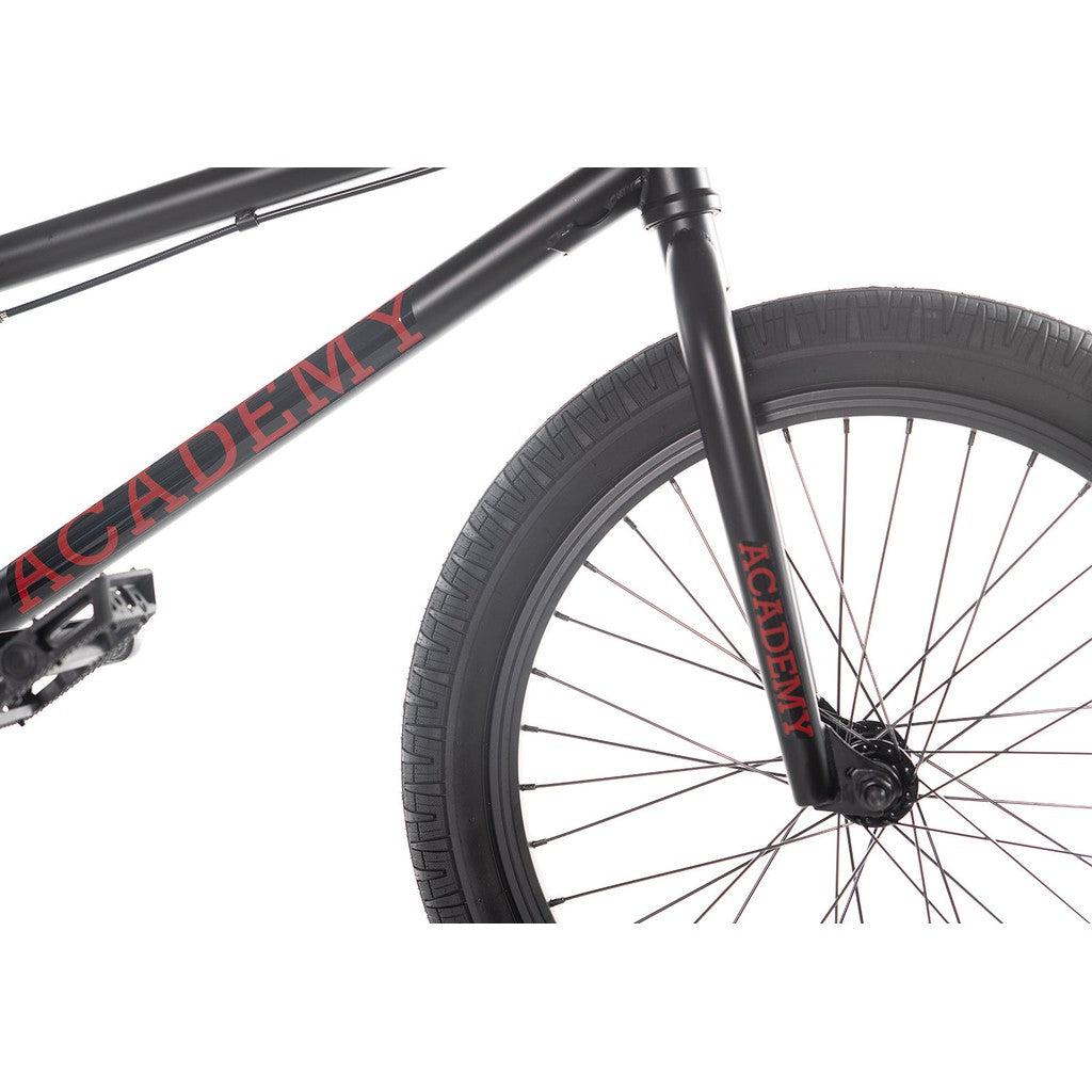 Close-up of a black BMX bike with "Academy" written in red on its chromoly frame and front fork. The focus is on the front section of the bike, highlighting the tire and a pedal from the Academy Desire 20 Inch Bike series.