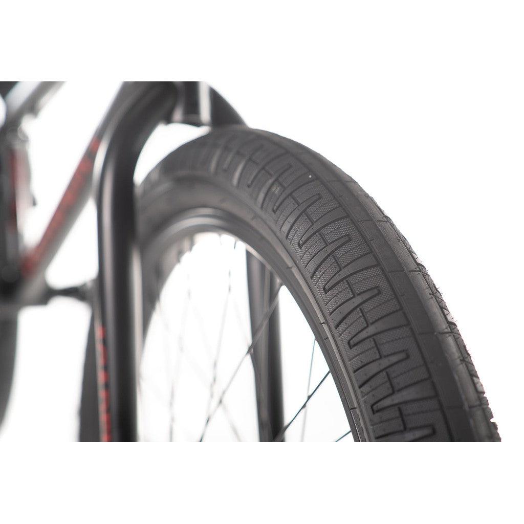 Close-up view of the front tire of a black Academy Desire 20 Inch Bike, showcasing its tread pattern and spokes against a white background, complemented by its full Chromoly frame and 21-inch top tube.