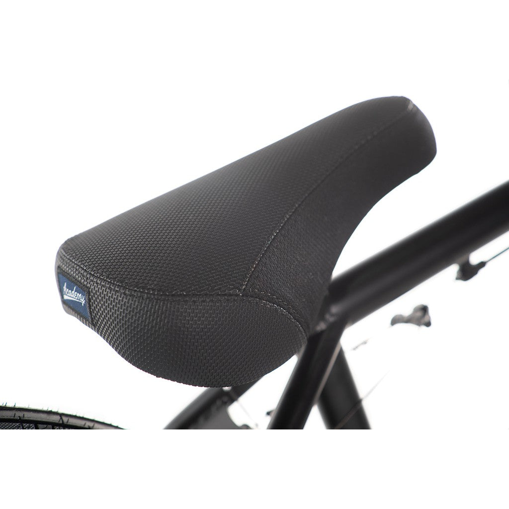 Close-up view of a black textured bicycle seat mounted on the full Chromoly frame of an Academy Desire 20 Inch Bike, featuring a small fabric tag on the side. The background is white.