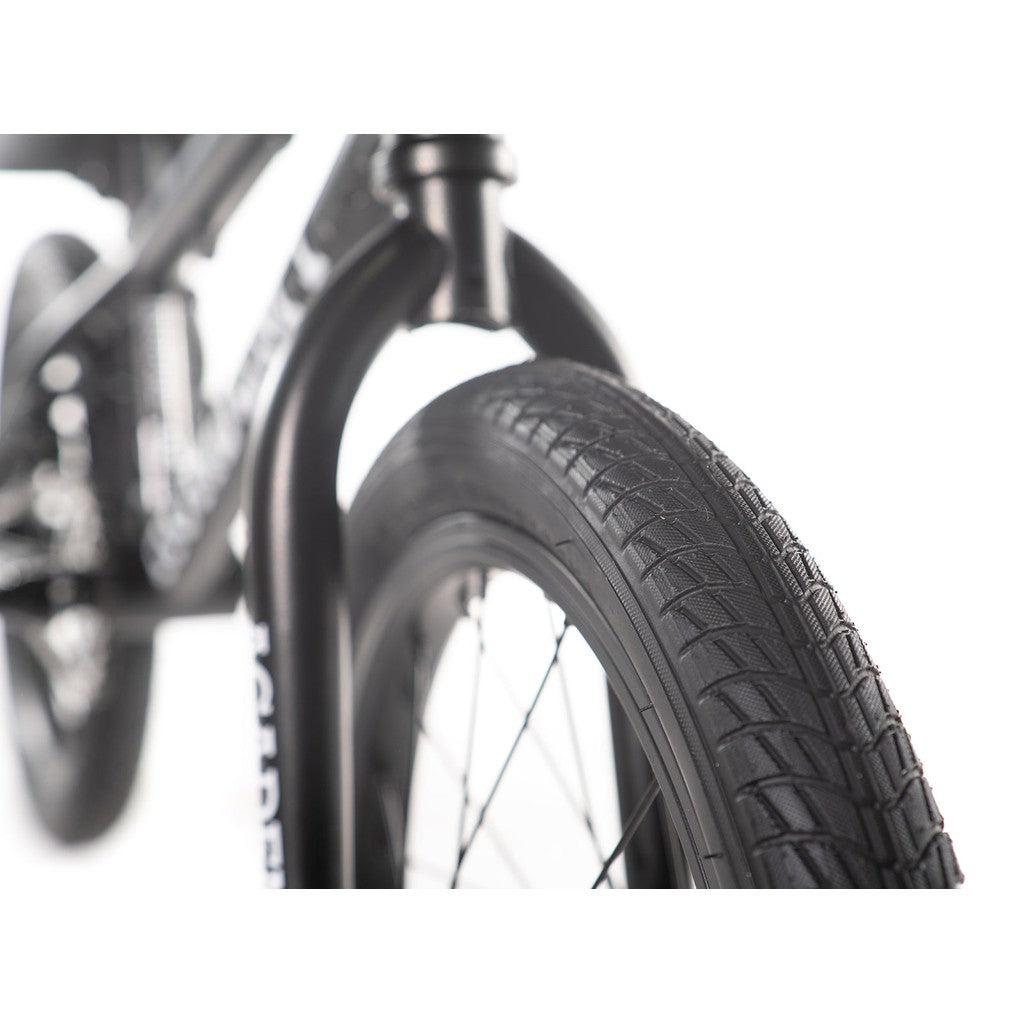 Close-up of a black bicycle tire with visible tread pattern, highlighting part of the sturdy CrMo frame on the Academy Inspire 16 Inch Bike in the background.