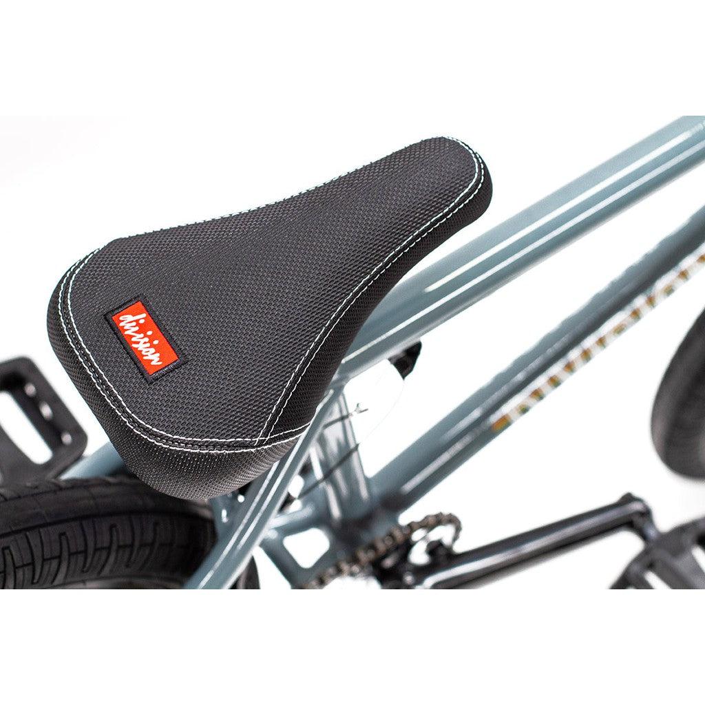 Close-up of the Division Fortiz 20 Inch Bike's black seat with white stitching and a red logo, mounted on its gray Chromoly frame BMX bike.