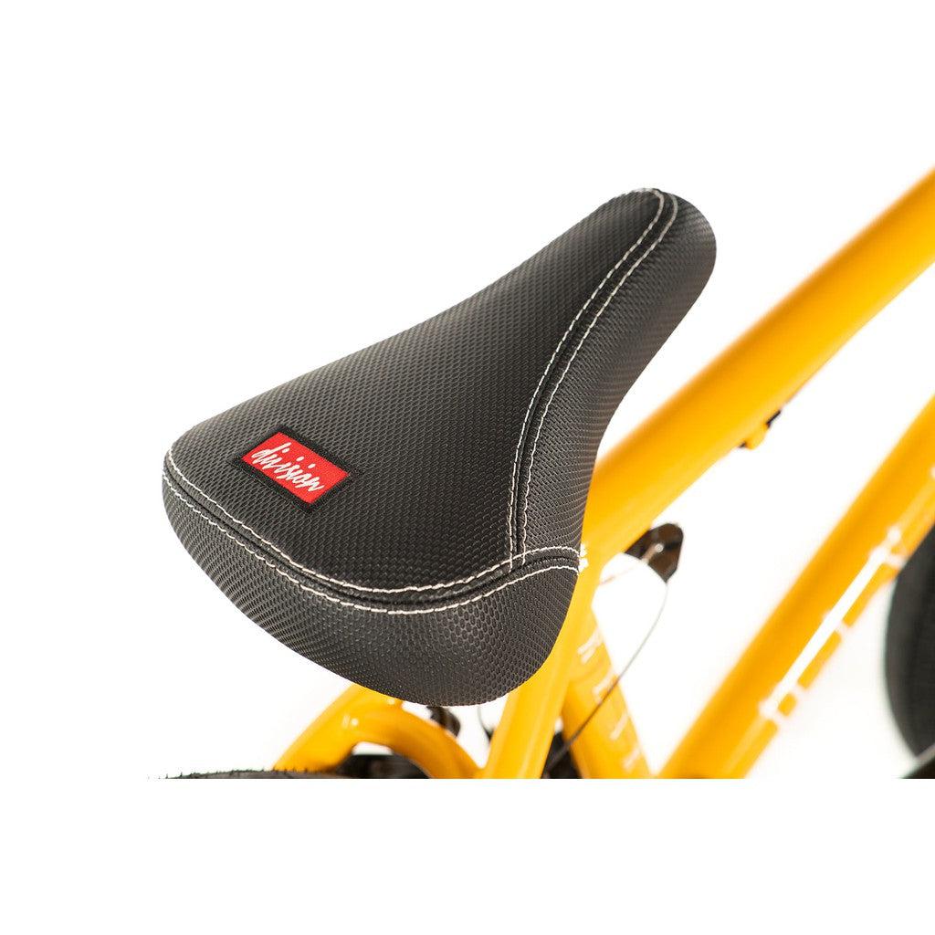 Close-up of a black bicycle seat with white stitching and a red label on a yellow Division Reark 20 Inch Bike.