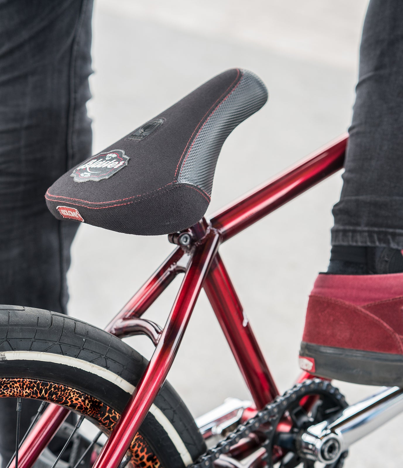 Close-up of a red bicycle featuring the Colony Blaster Fat Pivotal Seat, partially visible rear tire, and a person in red shoes standing nearby.