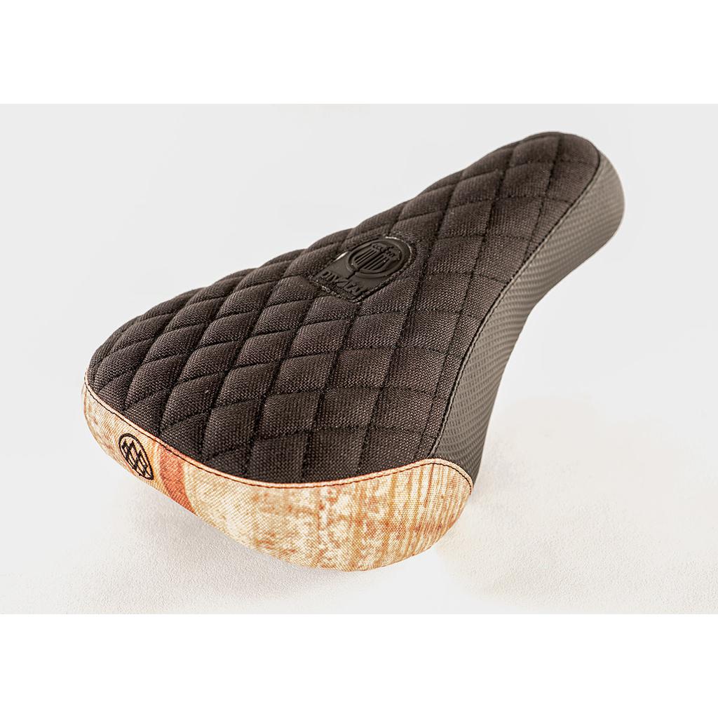 A black quilted bicycle seat with diamond stitching and a stitched emblem, accented by a brown bottom edge, reminiscent of the Colony Wallwork Pivotal Seat, photographed on a plain background.