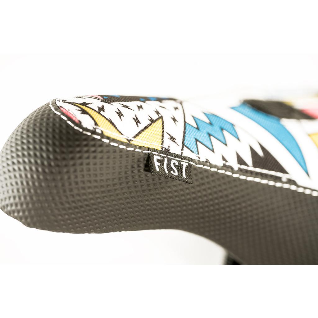 Close-up of the Colony X Fist Pivotal BMX seat with colorful geometric patterns and a small "FIST" tag. The edge features textured black fabric with extra grip and white stitching, akin to the durability of Fist Zig Zag gloves.