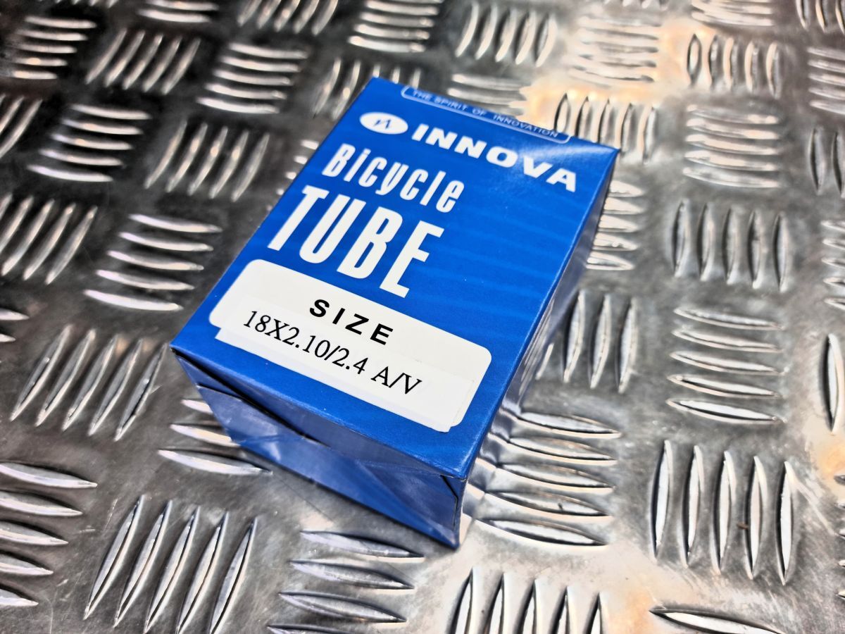 A blue box labeled "INNOVA Bicycle Tube Size 18x2.10/2.4 AV" sits on a metallic, textured surface, perfect for BMX riders looking for durable Colony Tube with a dependable Schrader valve system.