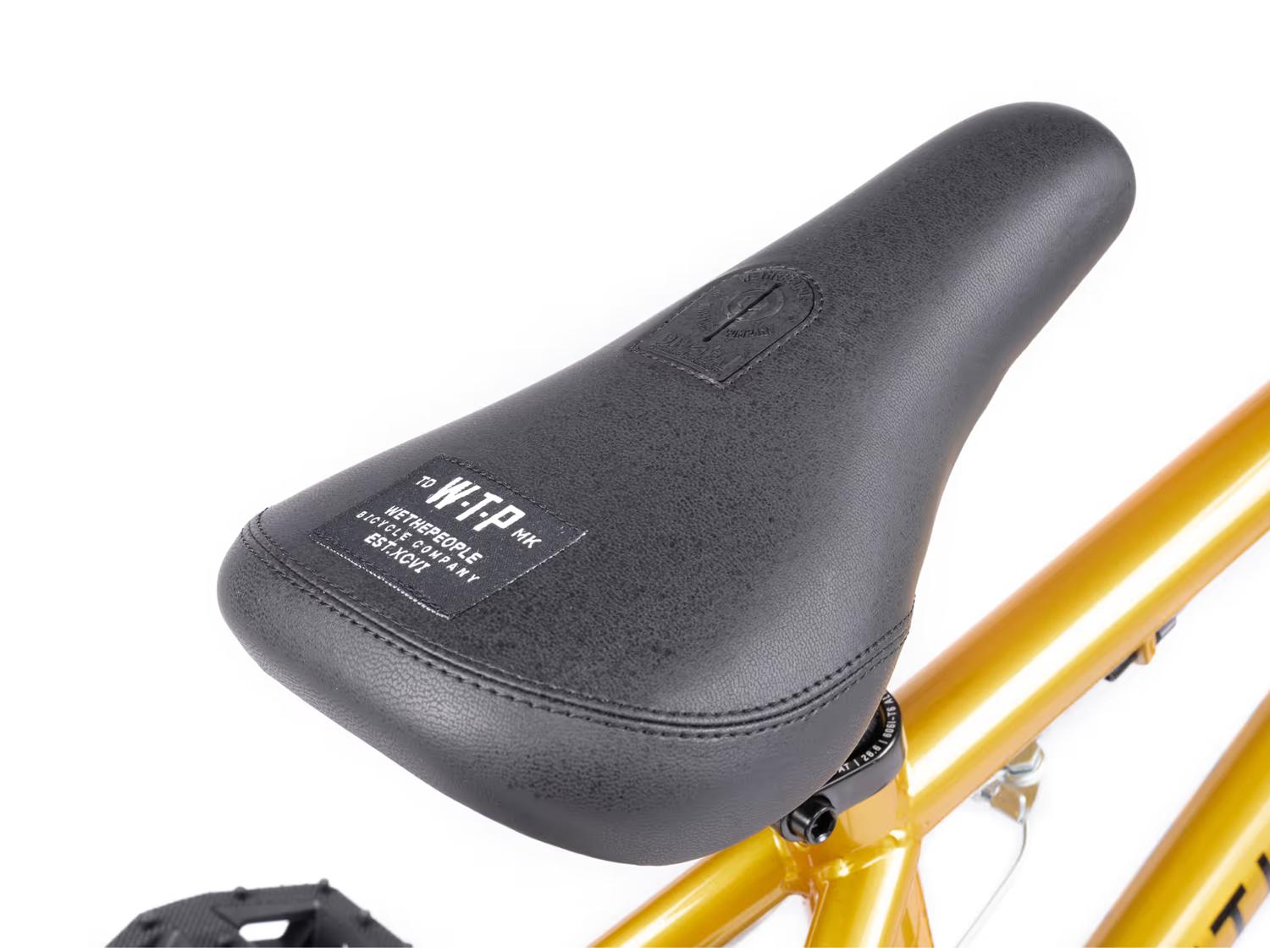 Close-up of a black seat with a fabric label on the Wethepeople Seed 16 Inch Bike, featuring a metallic gold frame, ideal for BMX shredders.