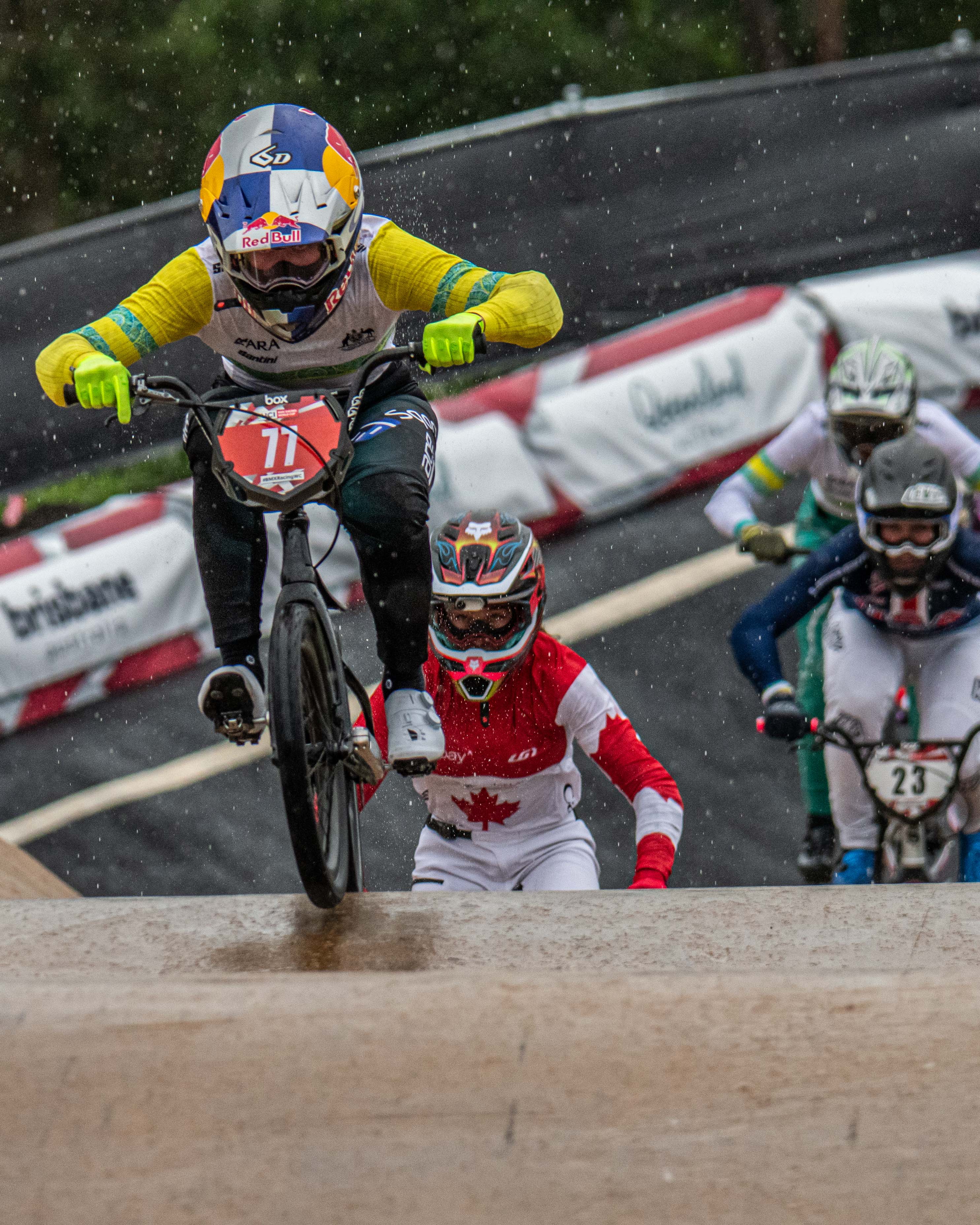 During the 2024 National BMX Championships in Perth, cyclists equipped with protective gear race their BMX bikes on a wet track. The lead rider, highlighted in the LUXBMX Race Photo Package, wears a distinctive helmet featuring a Red Bull logo. Rain pours down in the background, heightening the drama of this thrilling event.