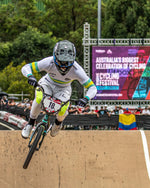 During the 2024 National BMX Championships in Perth, a BMX rider wearing a white and green uniform tackles a dirt ramp, framed by a prominent LUXBMX banner and an enthusiastic crowd snapping exciting moments with the LUXBMX Race Photo Package.