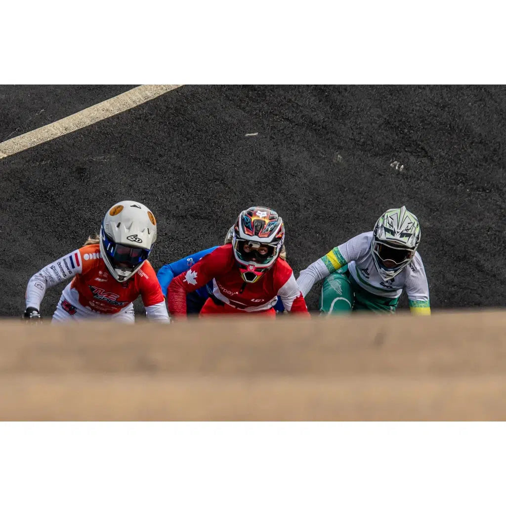 Three BMX riders in vibrant gear and helmets, seen from above, get ready at the starting line on a sloped track, eager to compete in the 2024 Perth Championships. Capture this exhilarating moment with high-quality photos from the LUXBMX Race Photo Package: 2024 National BMX Championships.