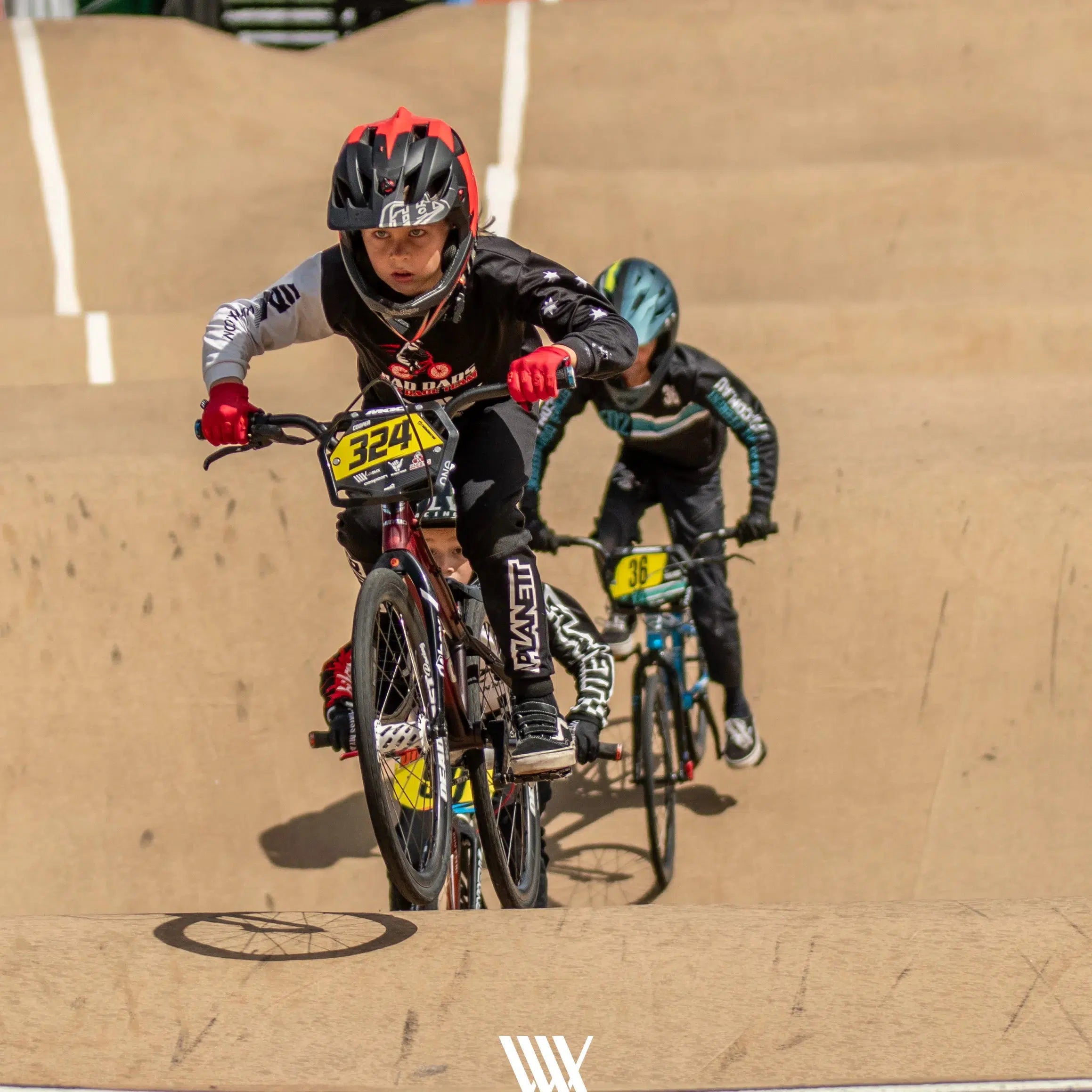 At the 2024 Perth Championships, two BMX riders race intensely on a dirt track, showcasing competitive spirit. The leading rider is equipped with a black helmet and red gloves, while the one following wears a sleek black and blue outfit. This thrilling scene is expertly captured in LUXBMX Race Photo Package: 2024 National BMX Championships, epitomizing the excitement of LUXBMX's signature events.