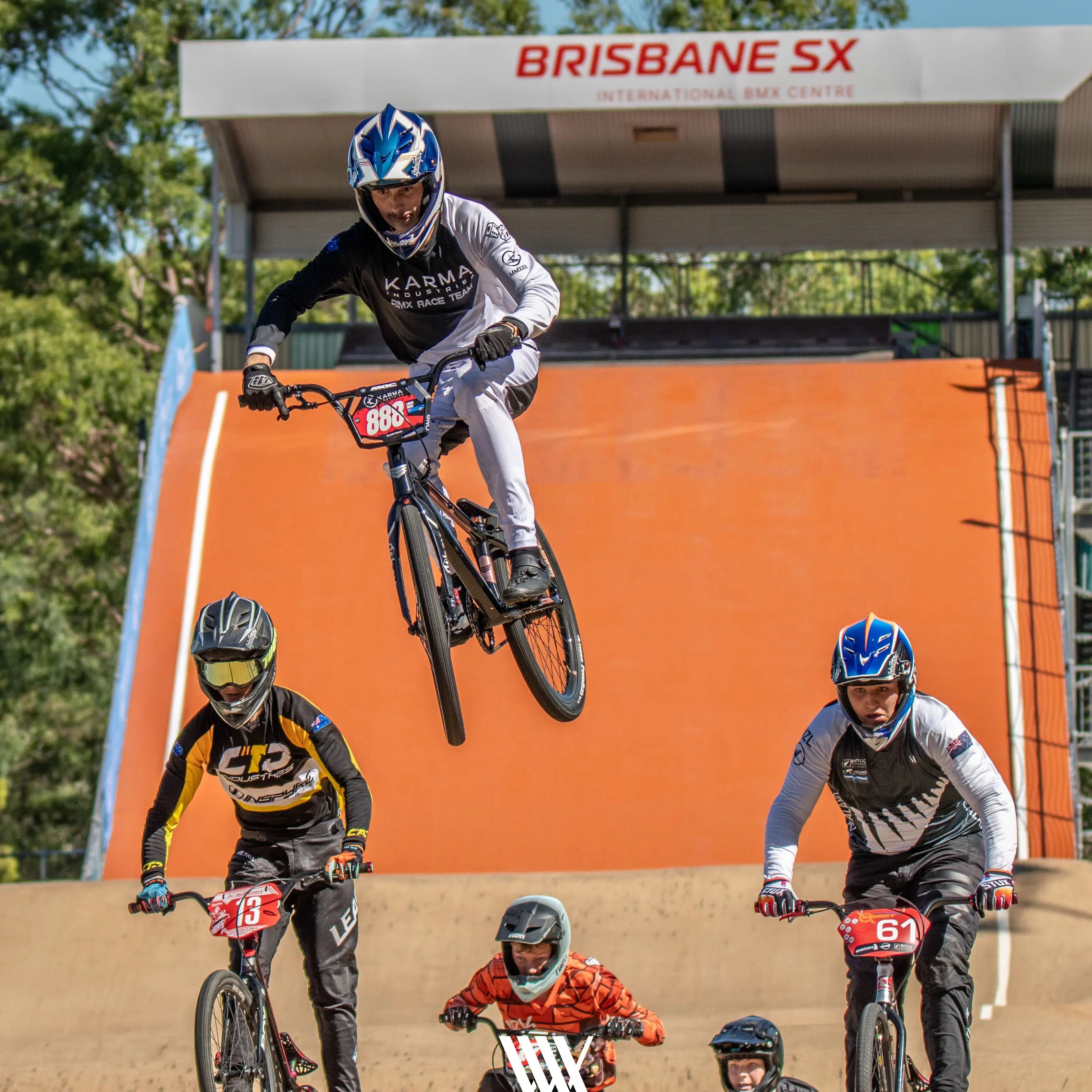 Cyclists race on BMX bikes at Brisbane SX, with one performer executing a jump in front of an orange ramp. Feel the thrill and excitement reminiscent of those captivating BMX race photos from the 2024 National BMX Championships, available in the LUXBMX Race Photo Package.