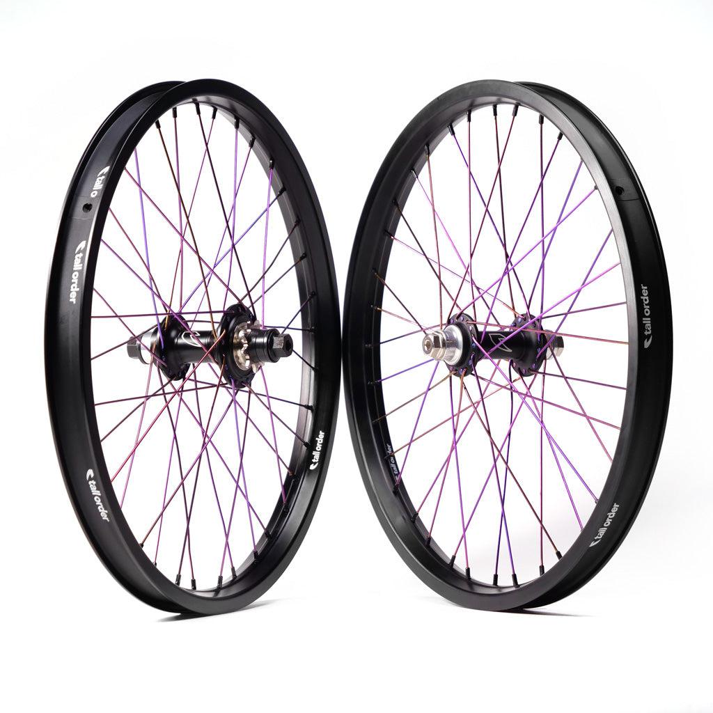 Two bicycle wheels are positioned upright facing each other. The spokes are colored in a mixture of purple and pink, and the rims and hubs are black. This custom-built wheel set features the Tall Order Drone x Air Custom Wheelset, branded with "tall order.