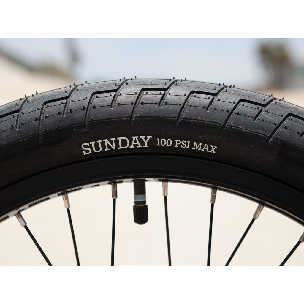 Close-up of a 20-inch Sunday Street Sweeper bike tire, highlighting the brand name "SUNDAY" and "100 PSI MAX" on the rubber. Designed by Jake Seeley, this model guarantees durability and performance, making it ideal for those who value quality craftsmanship.