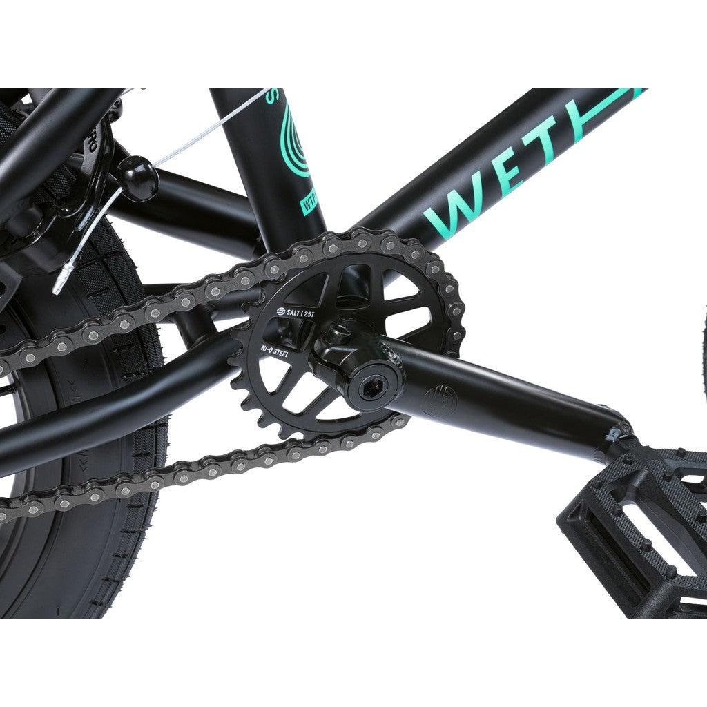 Close-up of the black Wethepeople Seed 16 Inch Bike with Salt Rookie Cranks, chain, and pedals, showing off "SALT" and "WETHEPEOPLE" branding in vibrant teal.