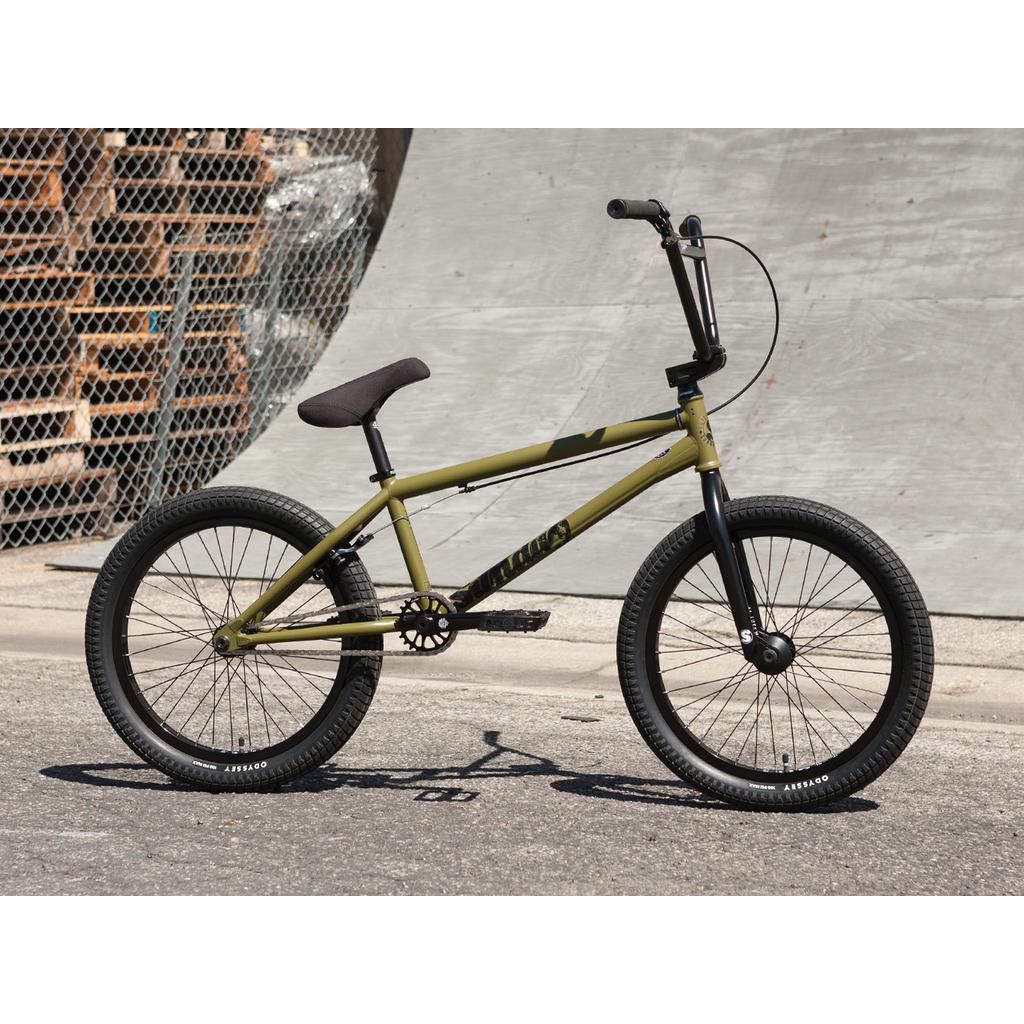 A Sunday Wave Length 20" (Gary Young) bike, in green and equipped with Odyssey Parts, features black handlebars and tires. It is parked on a paved surface near a chain-link fence and concrete wall.