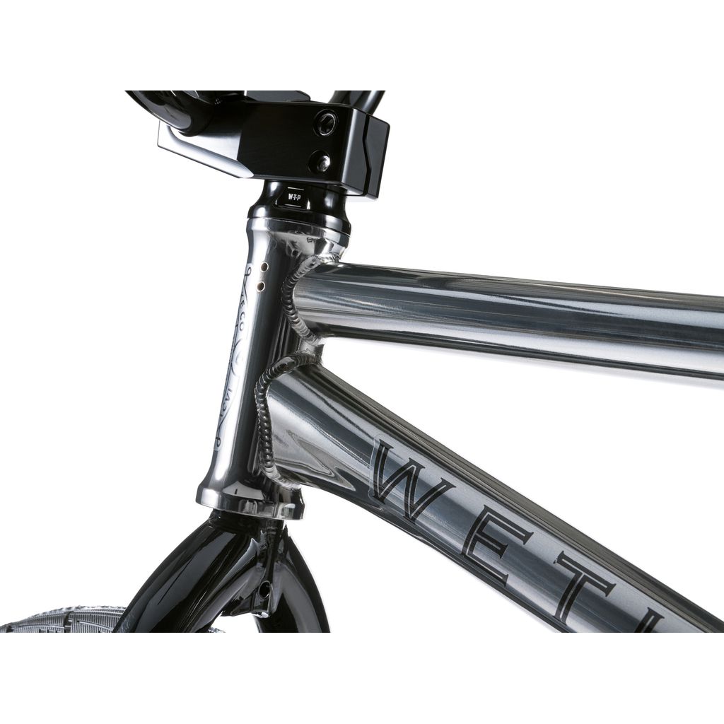 Close-up of a silver Wethepeople Envy 20 Inch BMX Bike, highlighting the intricate welds on its chromoly frame, complemented by a sleek black front tire against a crisp white background.