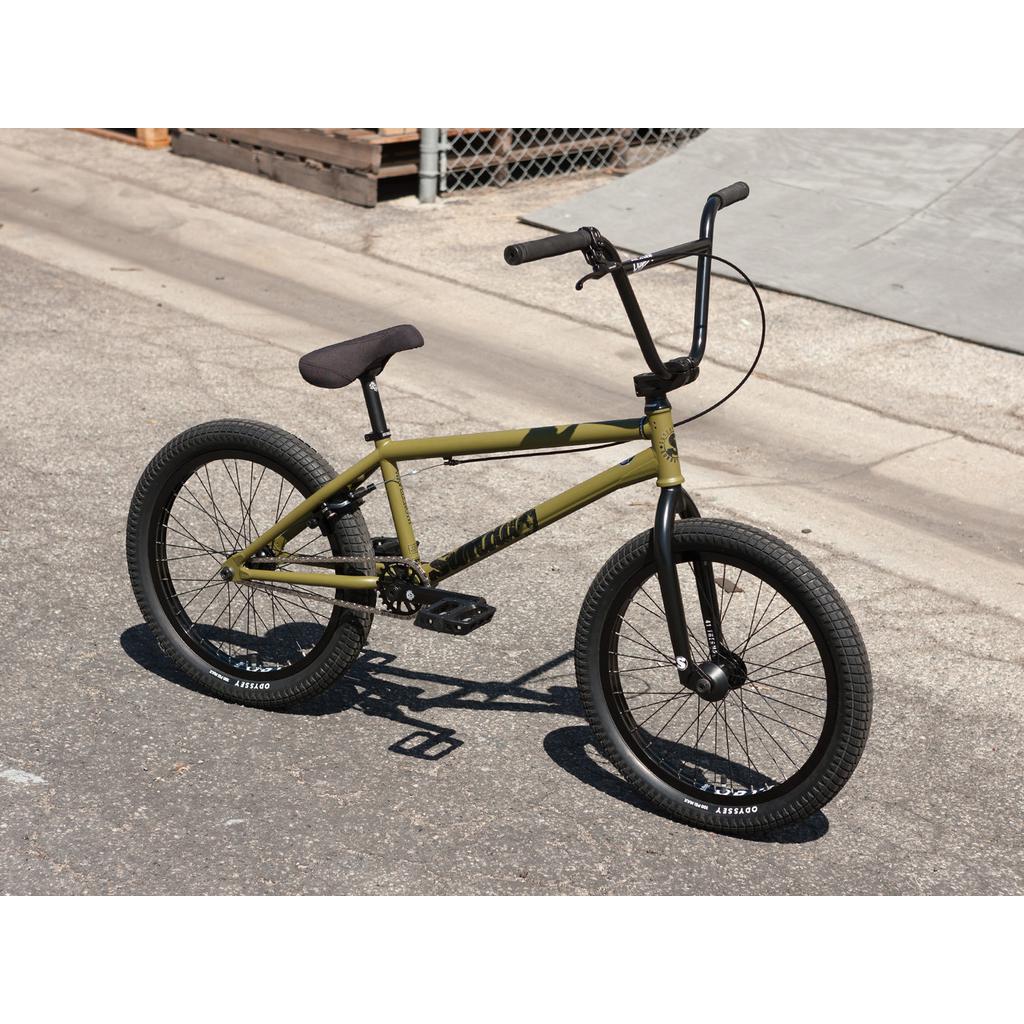 The Sunday Wave Length 20 Inch (Gary Young) BMX bike, featuring a green frame with black handlebars and tires, is parked on a paved surface near a chain-link fence, embodying the adventurous spirit of its namesake.