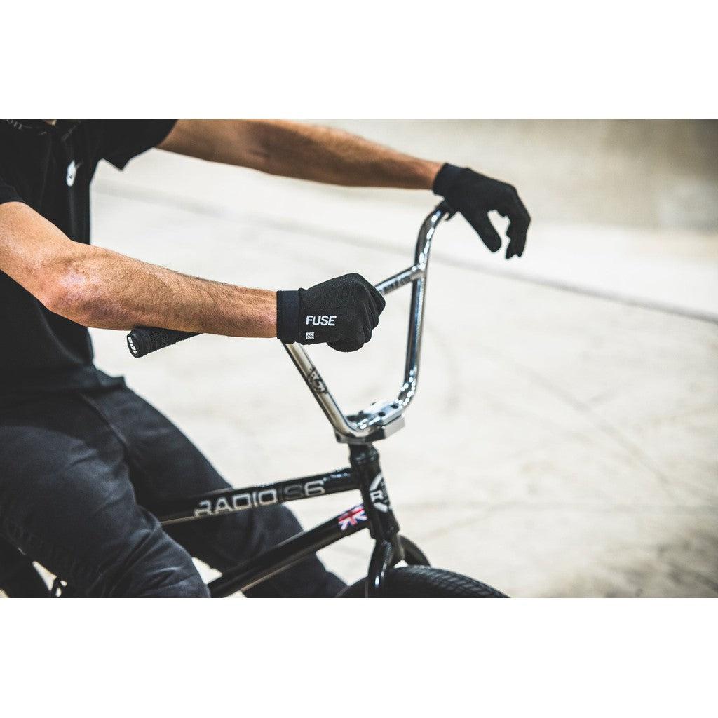 A person wearing Fuse Alpha Youth Gloves with reinforced thumbs grips the handlebars of a black BMX bike indoors.