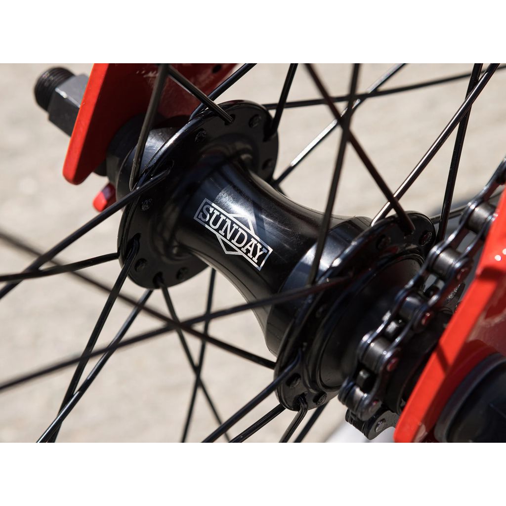 Close-up of a bicycle wheel hub labeled "Sunday," showcasing black spokes connected to a red frame. It's the hallmark of the sleek Sunday Primer 18 Inch Bike (2025), renowned for its Odyssey Springfield brakes.