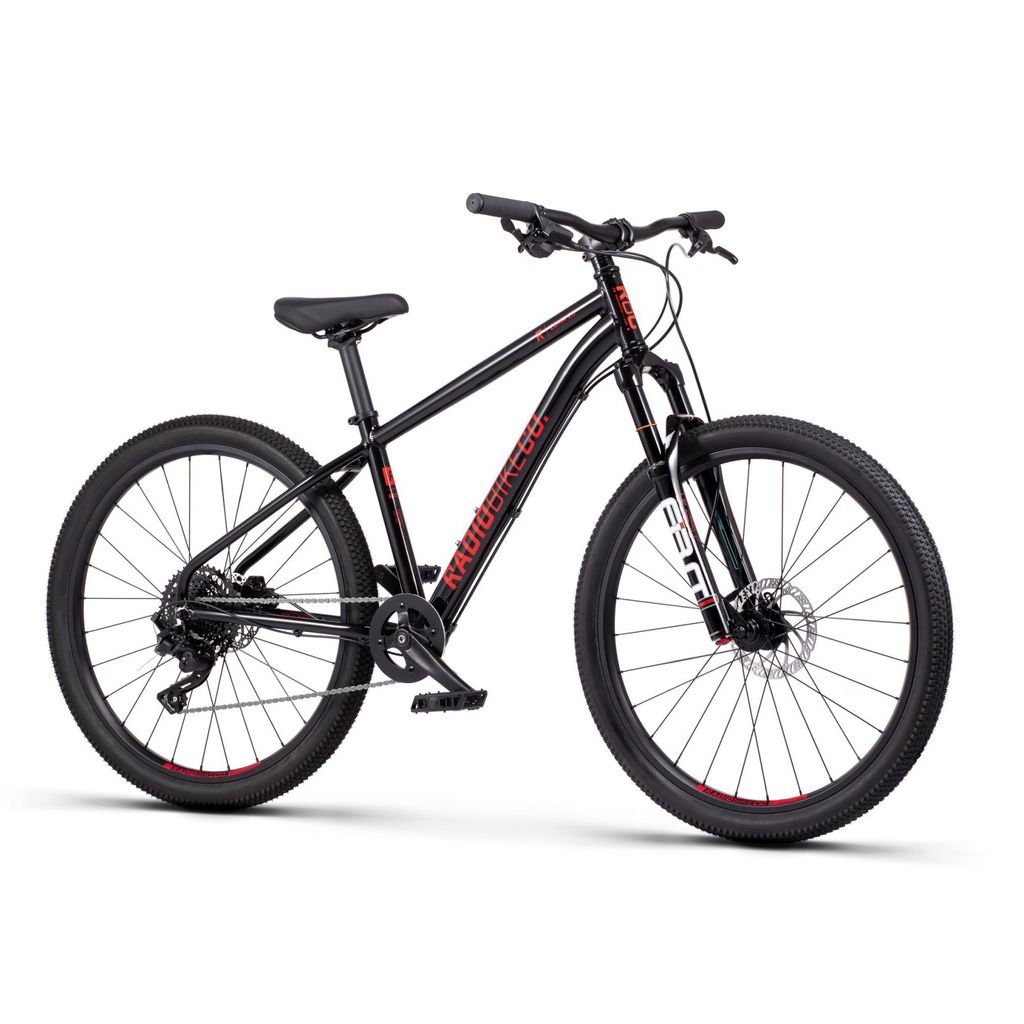 The Radio 26 Inch Zuma Sus Bike is a kid-sized MTB with a black lightweight alloy frame, featuring a red and white "Radon" logo, front suspension, wide knobby tires, disc brakes, and a flat handlebar.