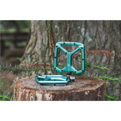 Two teal Funn Python V2 Alloy Pedals are on a tree stump in the forest—one upright and the other flat—showcasing their stability and performance amidst nature.