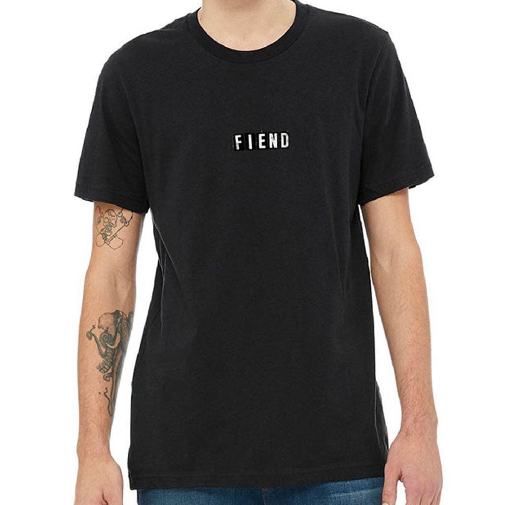 A person wearing a Fiend Ransom Vintage Shirt made of black, combed cotton with the word "FIEND" printed on the front, featuring tattoos on the left arm and blue jeans.