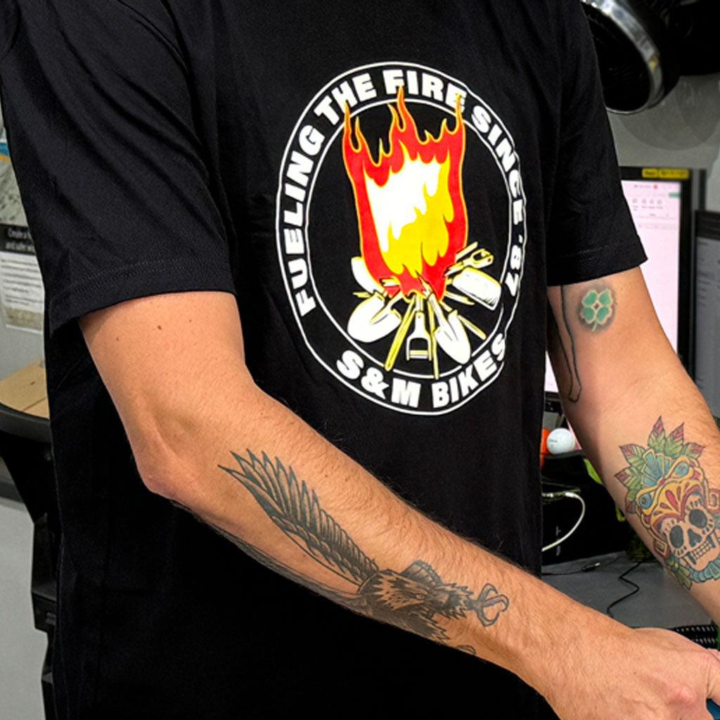 A person with tattoos on both forearms wears the S & M Fuel The Fire T-Shirt, which is black and features a logo with a burning match accompanied by the text "Fueling the fire since '87." This stylish tee is made of combed ring-spun cotton, ensuring comfort and durability.