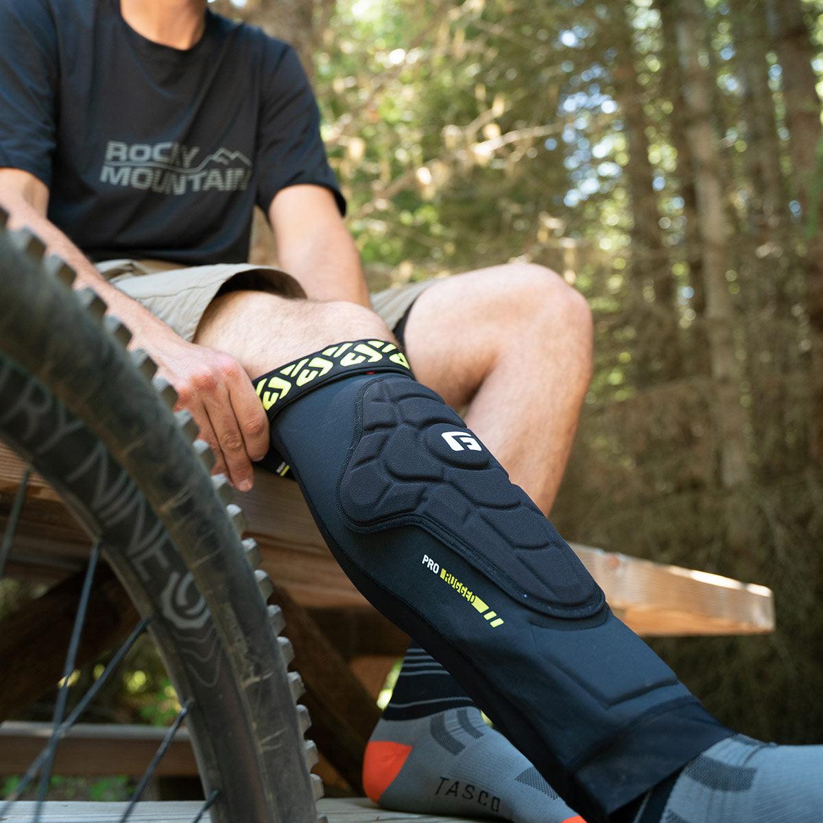 In a serene forest setting, a person sits on a bench carefully adjusting their G-Form Pro-Rugged 2 Knee Pads (Pair) next to a bicycle wheel, readying for an exhilarating enduro riding adventure.