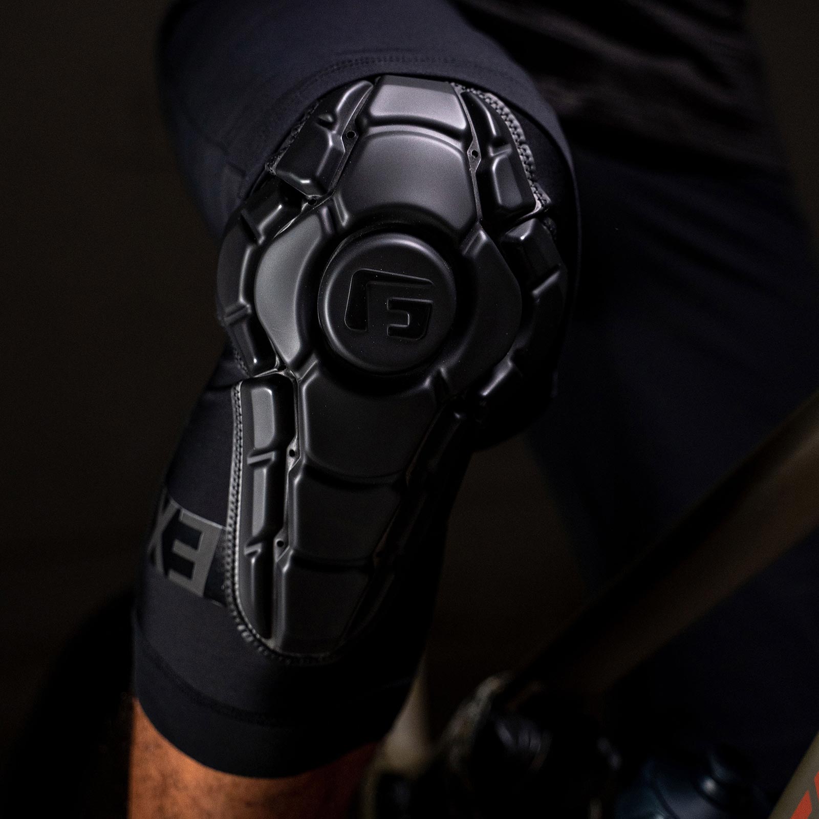 A person sits on a bicycle, wearing G-Form Pro-X3 Knee Guards with a textured surface featuring SmartFlex protection.