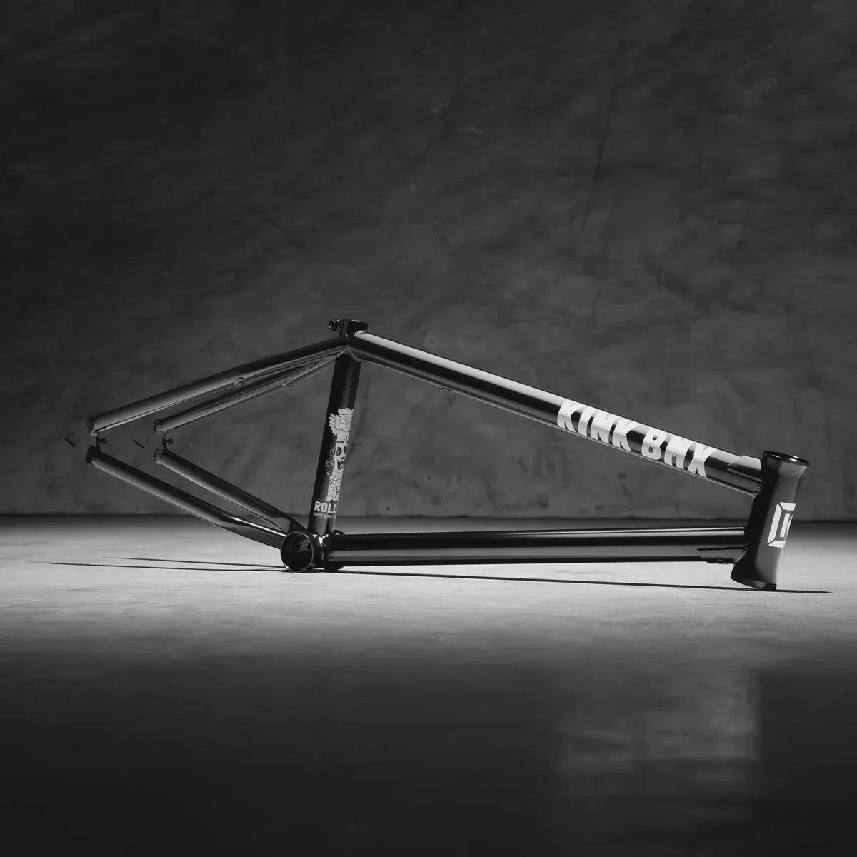 The Kink Roll Up Frame, a black bicycle frame with white text, rests on its side against a dark textured background. Its modern street geometry enhances the sleek design while ensuring durability for urban adventures.