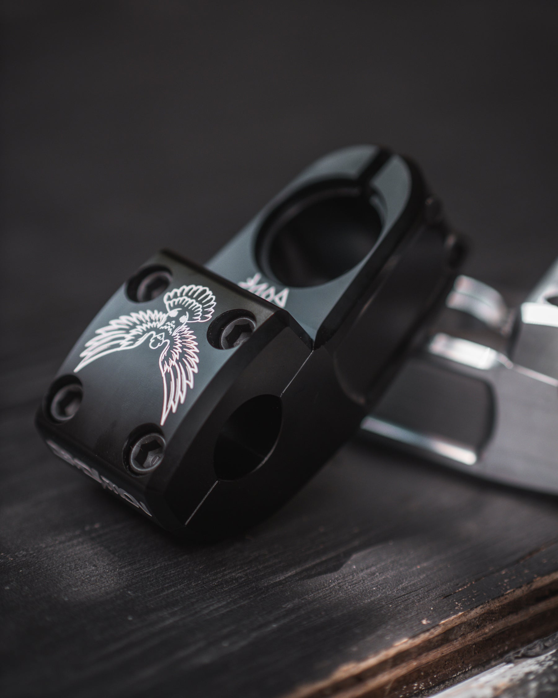 A close-up of the black metal Demolition Josh Dove Stem, featuring a white winged emblem design, rests elegantly on a dark wooden surface.