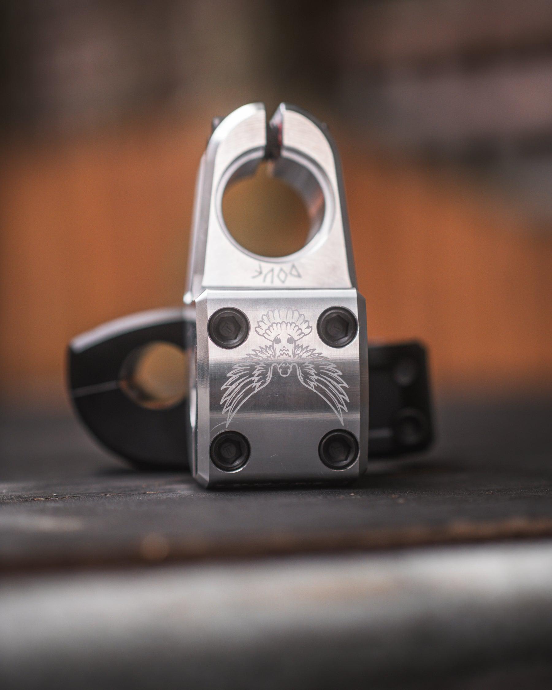 Close-up of the Demolition Josh Dove Stem with a stylized eagle engraving. Four screws ensure durability, while the blurred background in warm tones highlights its intricate craftsmanship.