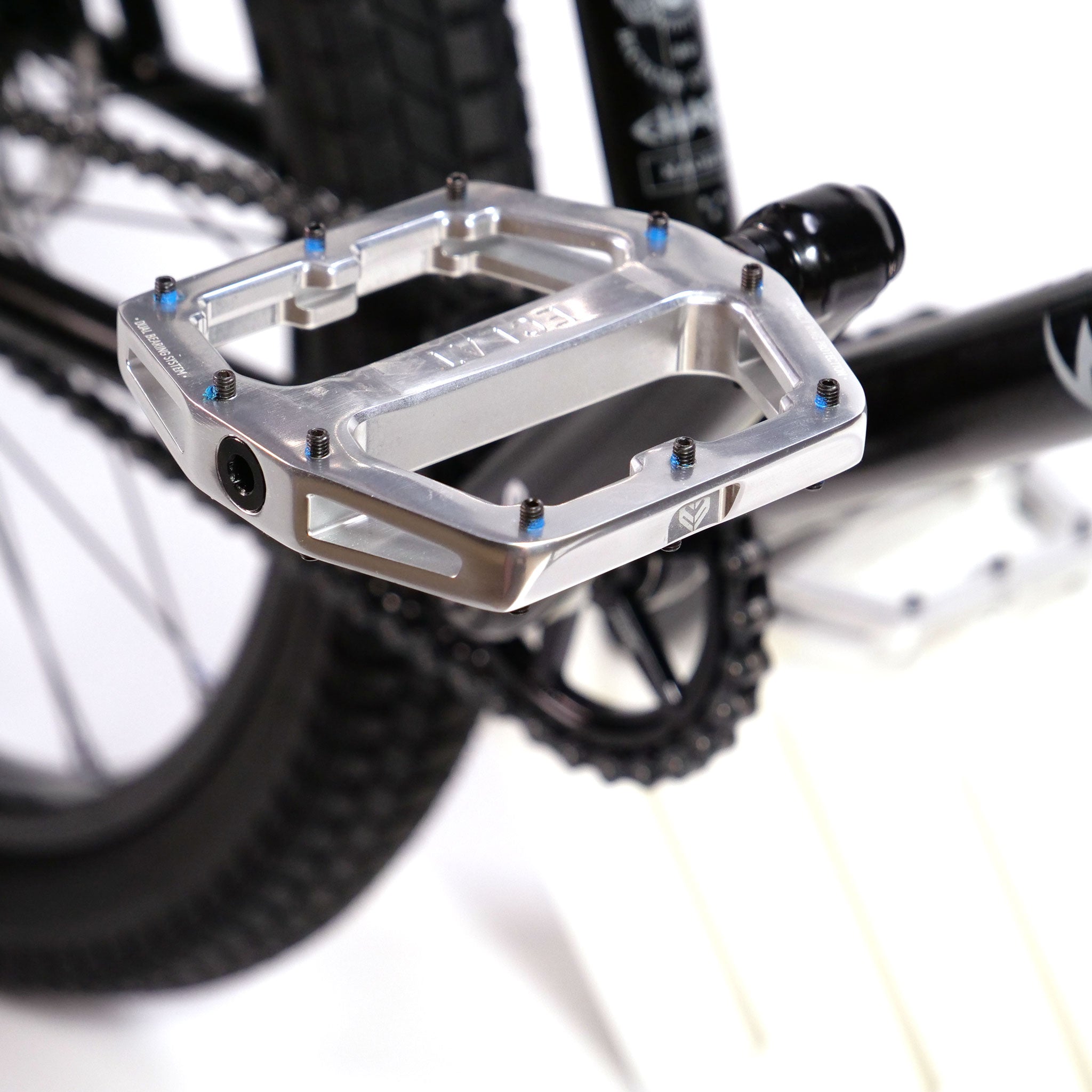 Close-up of an Eclat Exile silver bicycle pedal attached to the crank arm, with a part of the rear wheel and chain visible in the background, perfect for a Wethepeople Chaos Machine 22 Inch Custom Bike.