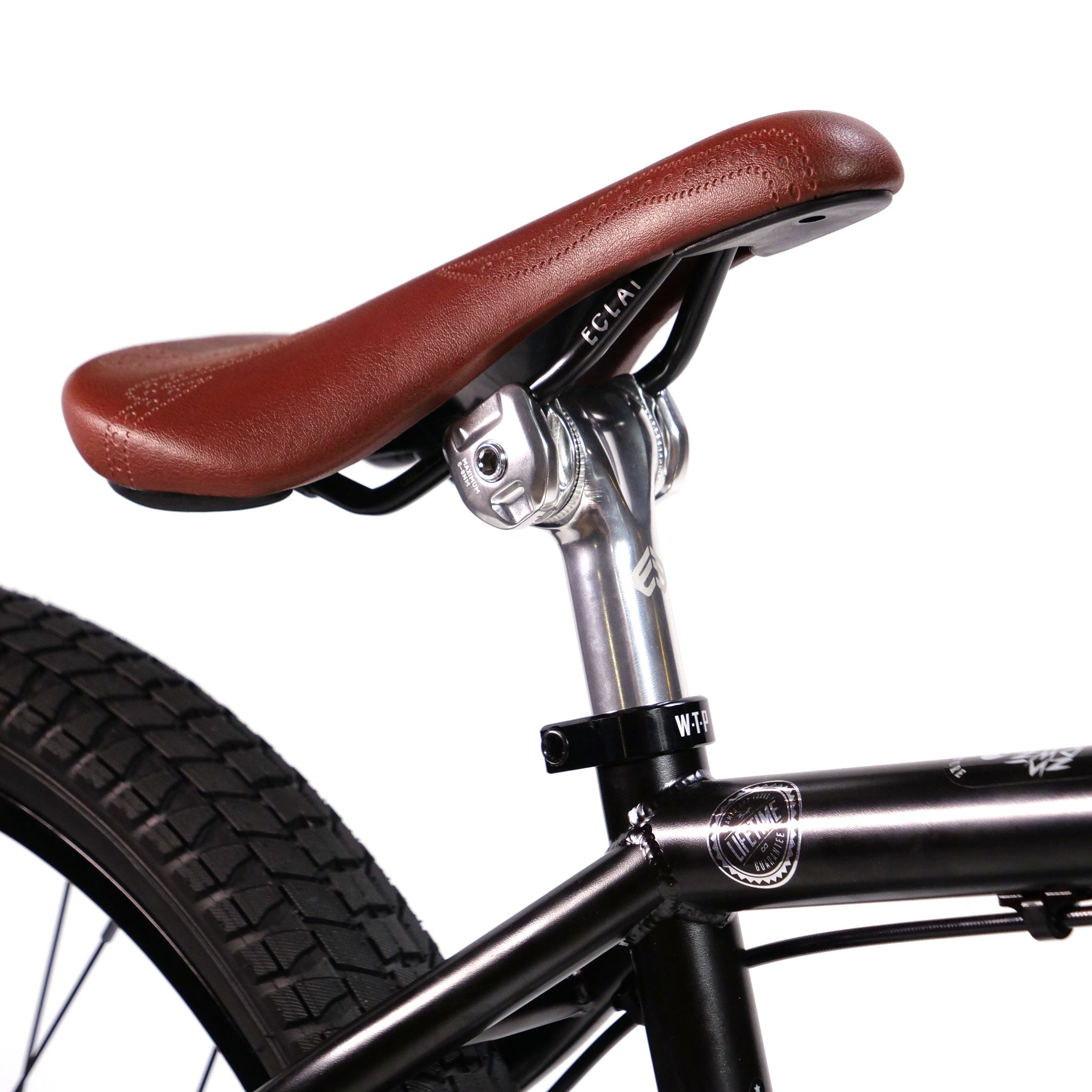 Close-up of a brown bicycle seat on a black frame, part of the Wethepeople Chaos Machine 22 Inch Custom Bike. The image shows the seat post, clamp, and part of the rear wheel with a textured tire.