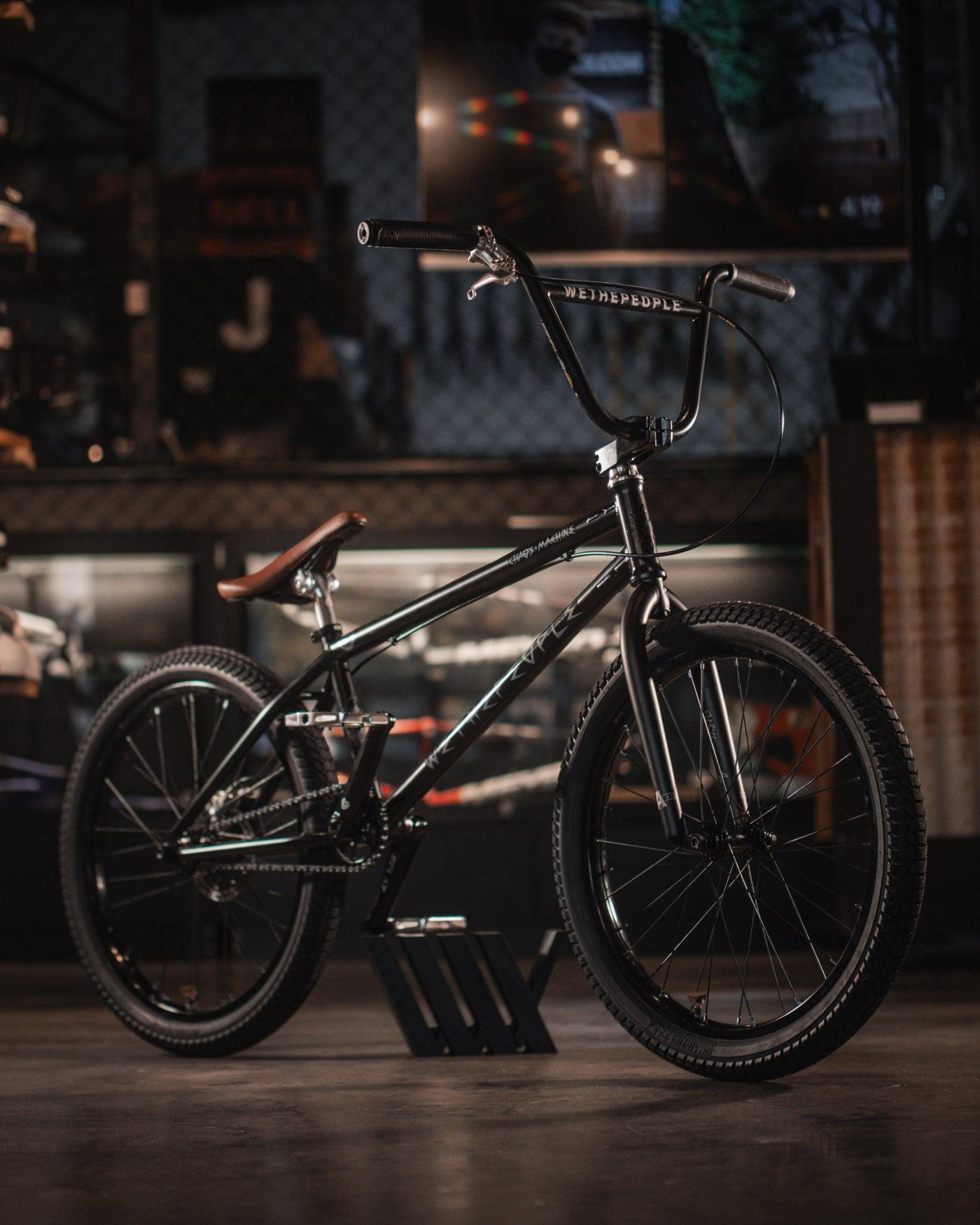 A black BMX bike with "WeThePeople" branding, also known as the Wethepeople Chaos Machine 22 Inch Custom Bike, is placed on a stand in a dimly lit indoor setting. The bike features thick tires, a brown leather saddle, and a clean chain.