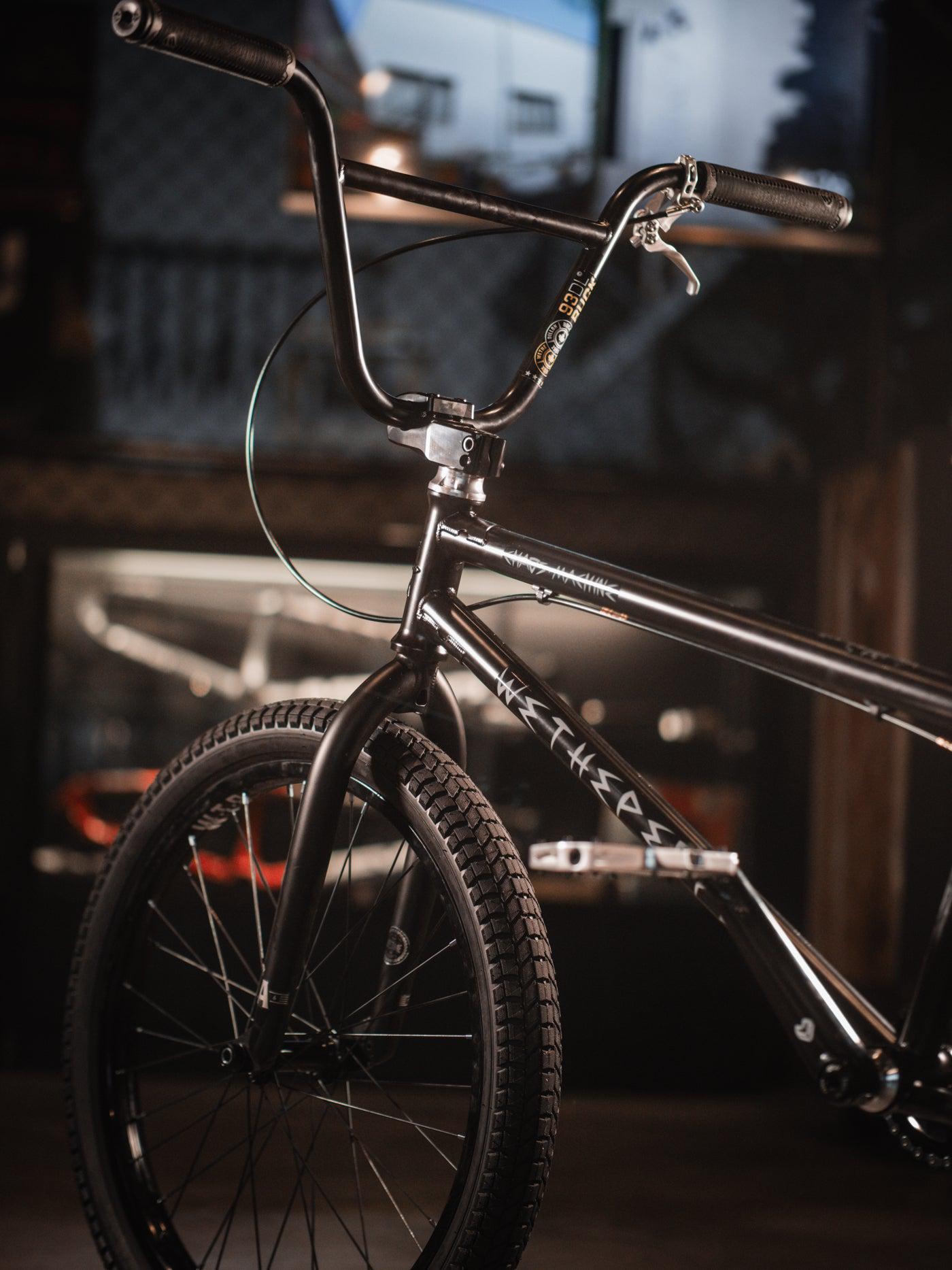 A close-up of a BMX bike in a dimly lit environment, showcasing the handlebars and front tire, highlighted by the sleek design of the Wethepeople Chaos Machine 22 Inch Custom Bike.