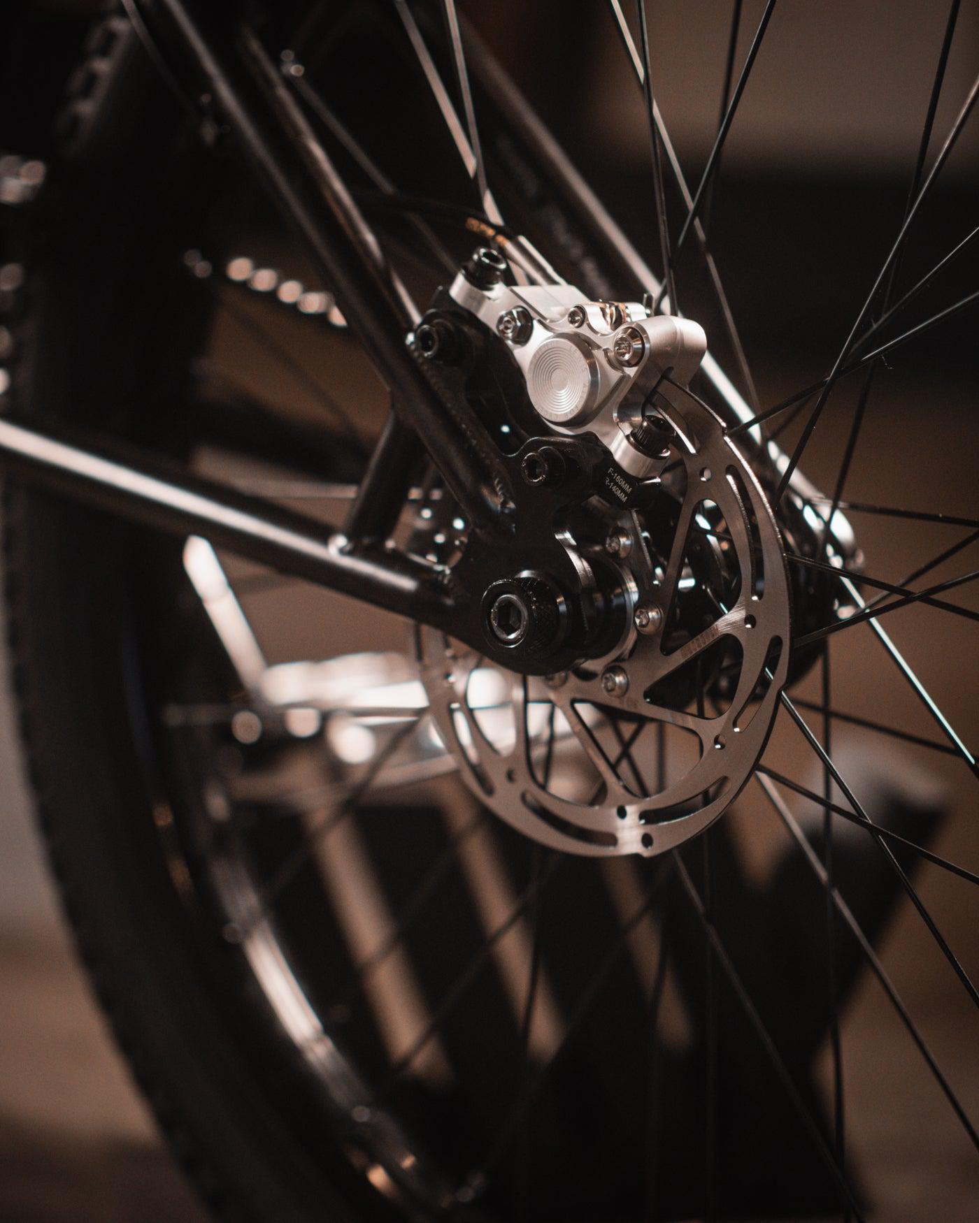 Close-up of a bicycle wheel focusing on the disc brake mechanism, showcasing the intricate details of the brake caliper and rotor, as seen on the Wethepeople Chaos Machine 22 Inch Custom Bike.