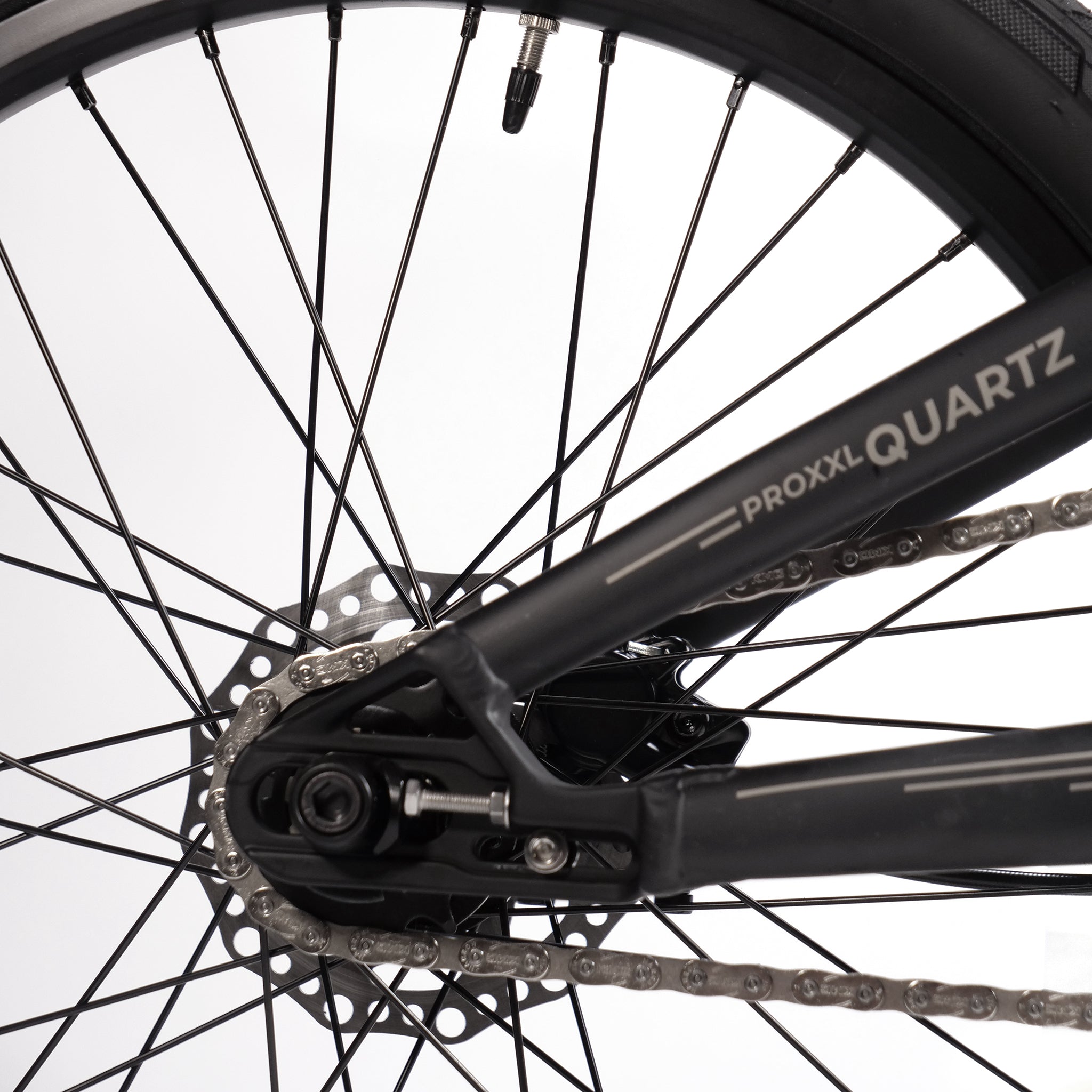 Close-up of the rear wheel of the Radio Raceline Quartz Pro L Bike 2025, showcasing the spokes, chain, and part of its lightweight hydro-formed frame with "PROXXL QUARTZ" branding. The background is white.