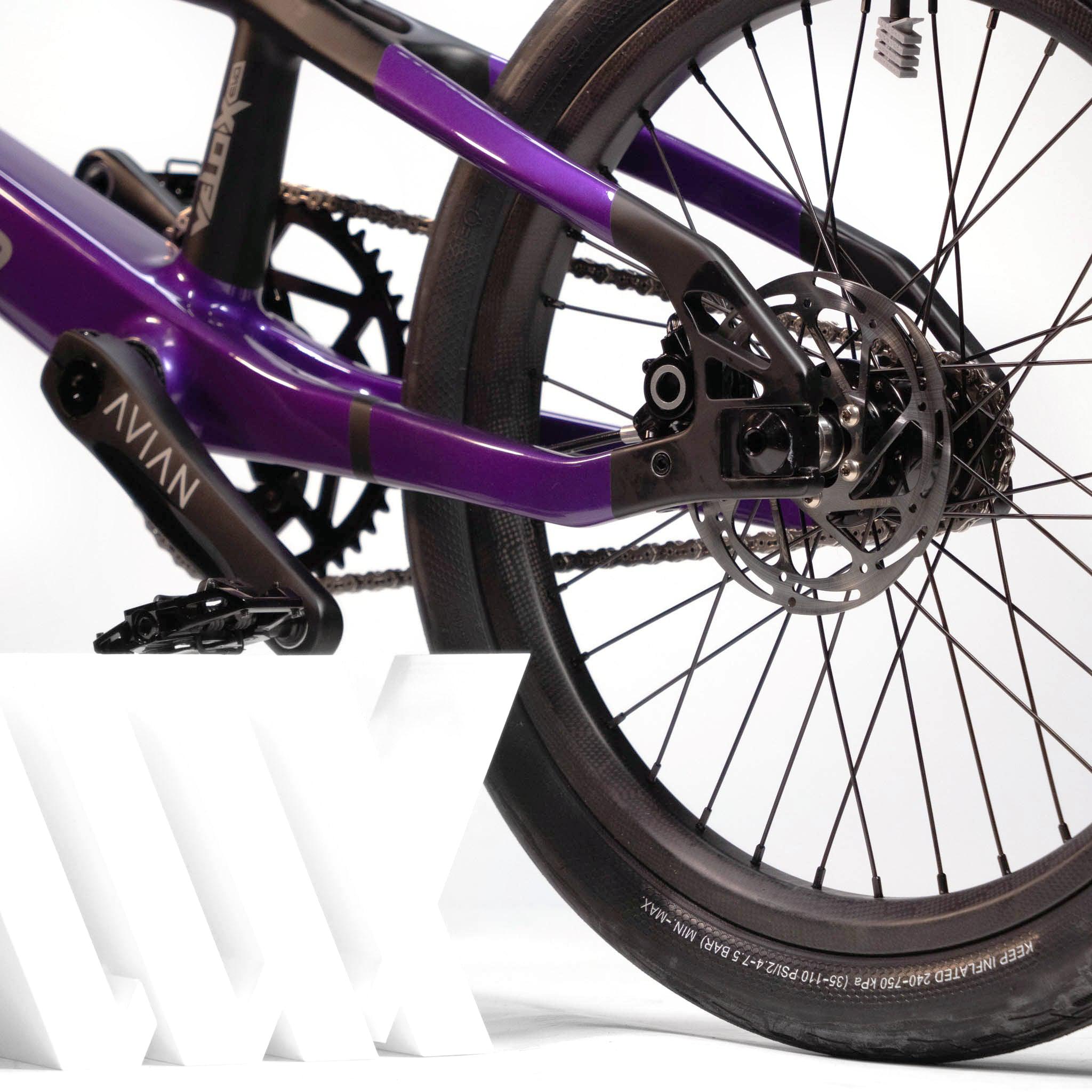 A close-up view of the Speedco Velox Evo Pro XXXL Custom Bike's rear wheel and brake system, highlighting the black disc brake, tire, and part of the frame. The background is white with a small portion of a logo visible. This custom race bike boasts premium Avian gear for optimal performance.