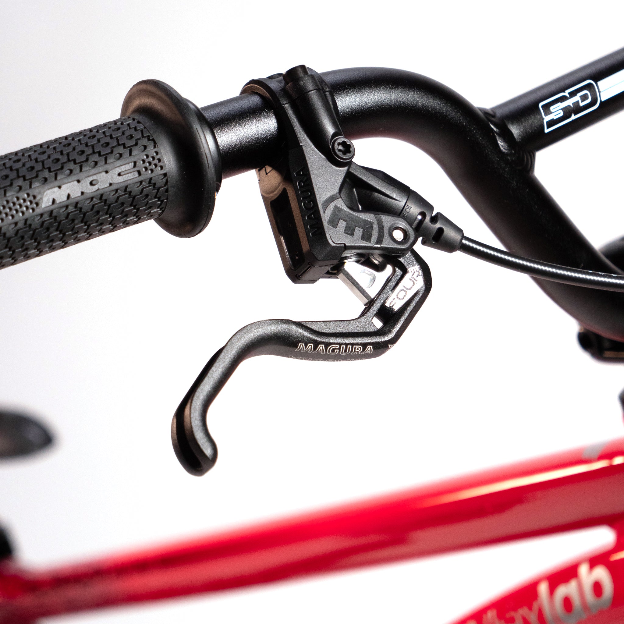 Close-up image of a black bicycle handlebar with a Magura brake lever on the Meybo Holeshot Junior Custom Bike. The handlebar features a textured grip and connects to the vibrant red frame outfitted with BOX carbon forks, highlighting the precision often seen in custom race bikes.