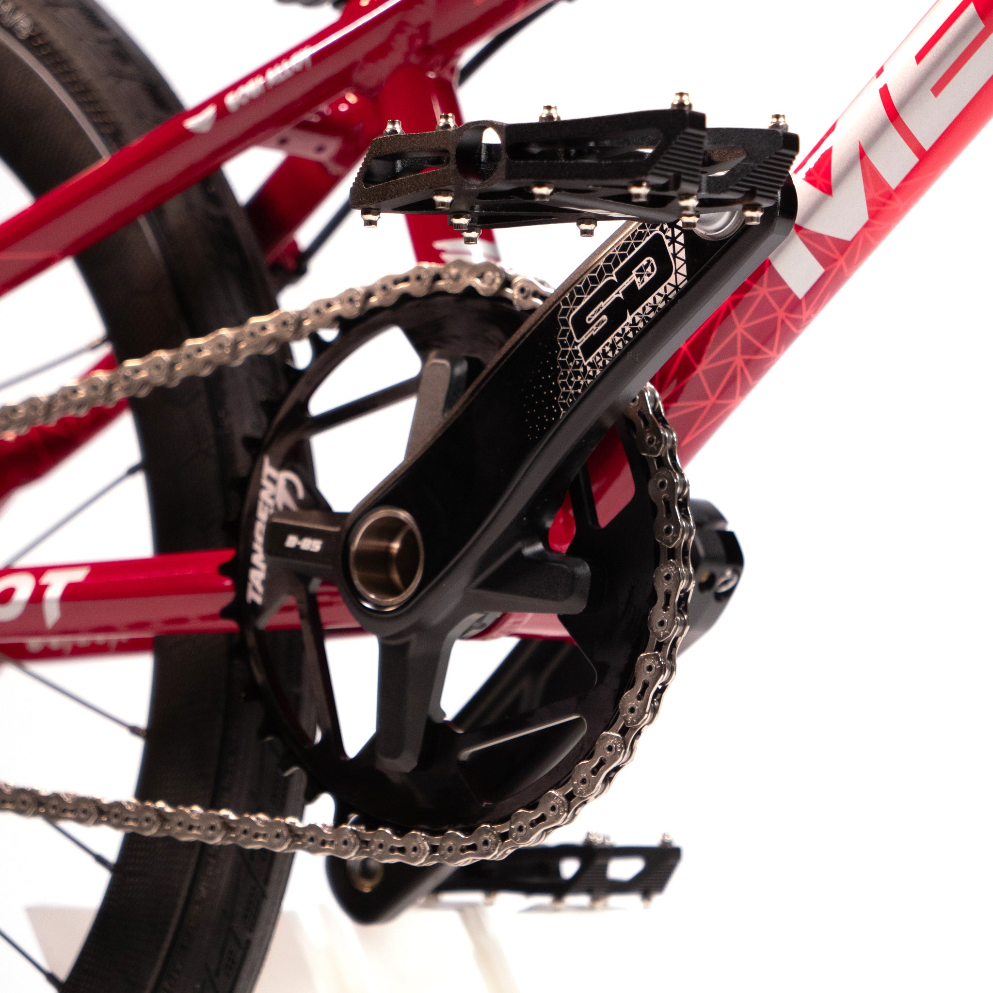 Close-up of a red bicycle's crankset and chain mechanism. The chain runs through a black pedal crank, while the rear wheel is partially visible in the background, showcasing the precision typical of custom race bikes like the Meybo Holeshot Junior Custom Bike.