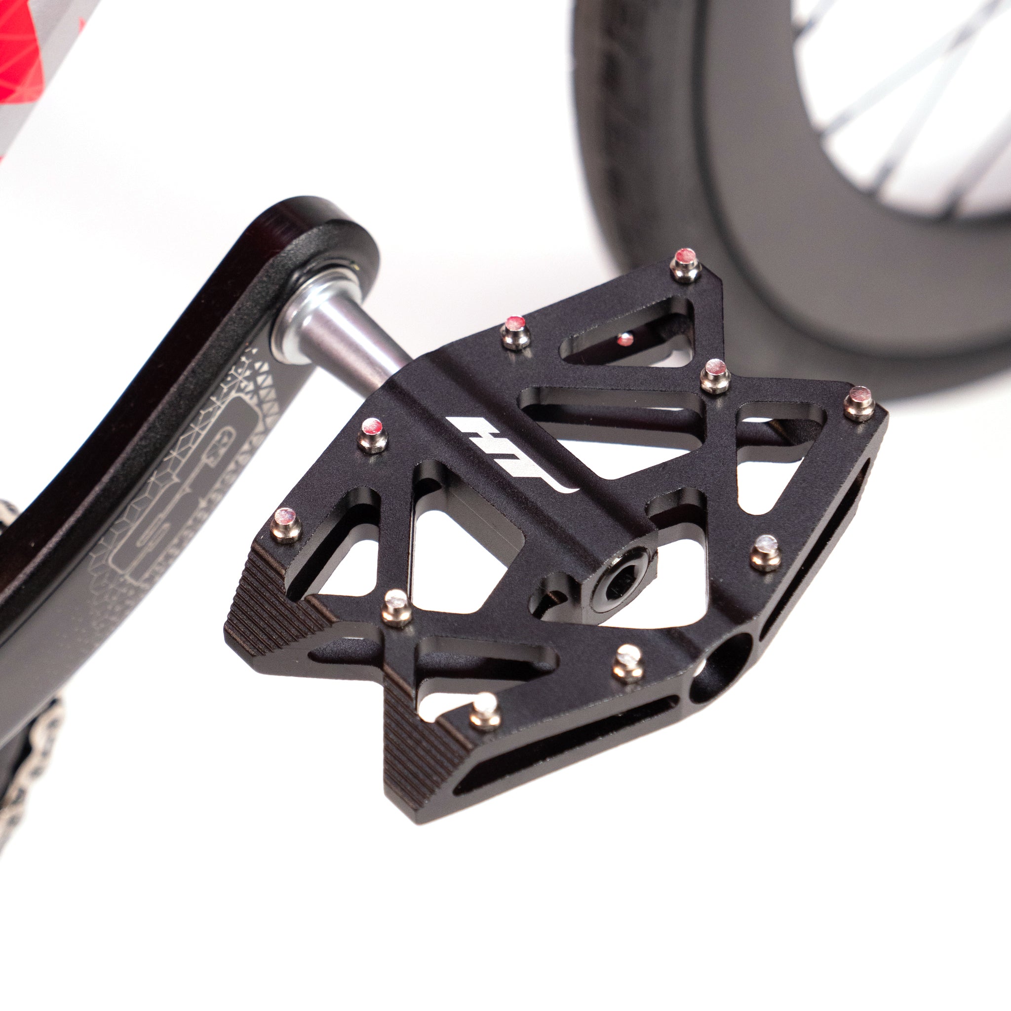 Close-up view of a black bicycle pedal attached to a crank arm, featuring a flat platform and metal pins for grip. In the background, you can see part of the wheel on the Meybo Holeshot Junior Custom Bike, highlighting its status as an excellent race bike.