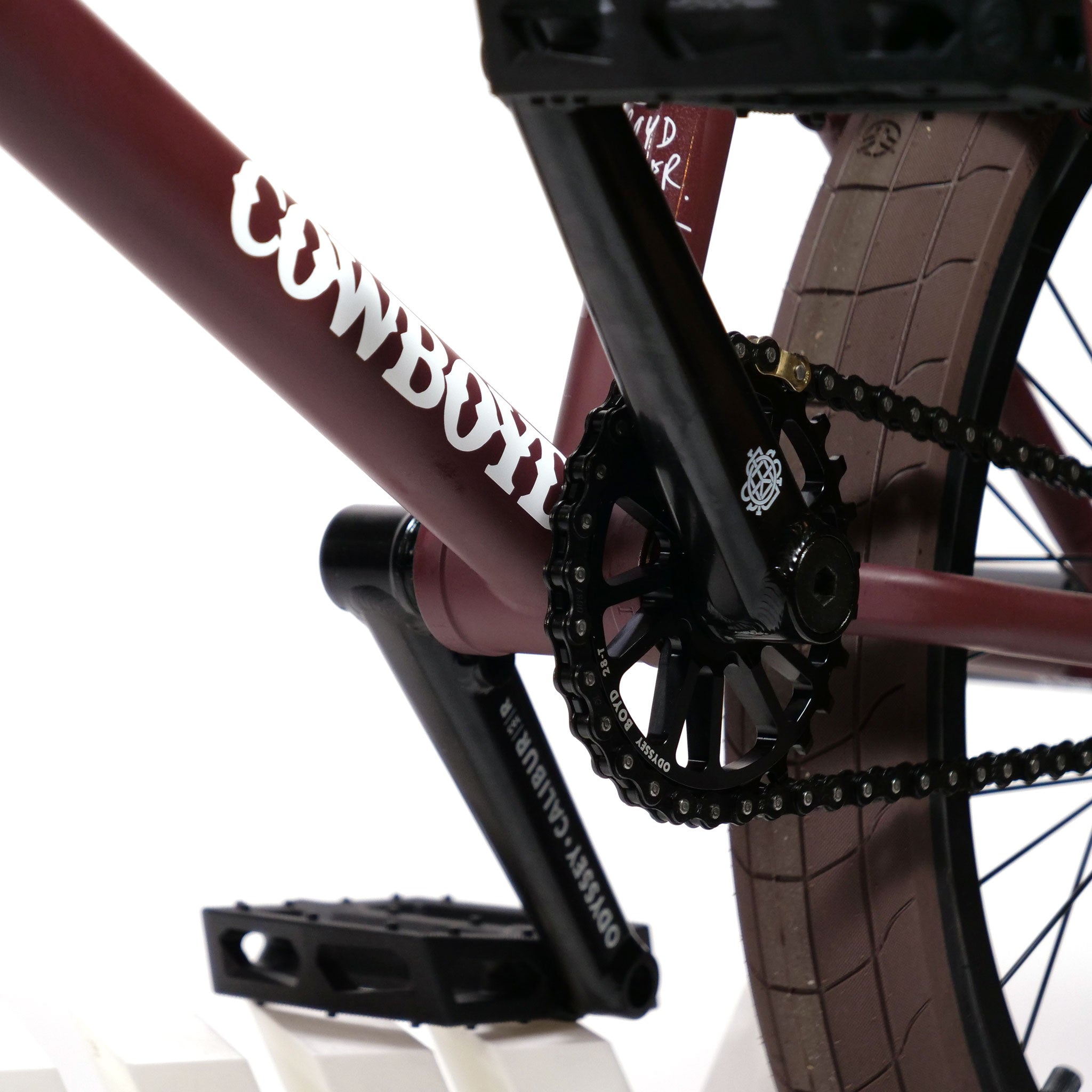 A detailed look at the frame of the Federal CowBoyd Custom 20 Inch Bike reveals a maroon-colored structure emblazoned with "COWBOYD," complemented by black pedals, a chainring, and a chain. In the background, a brown tire completes the streamlined design of this modern cycling masterpiece.