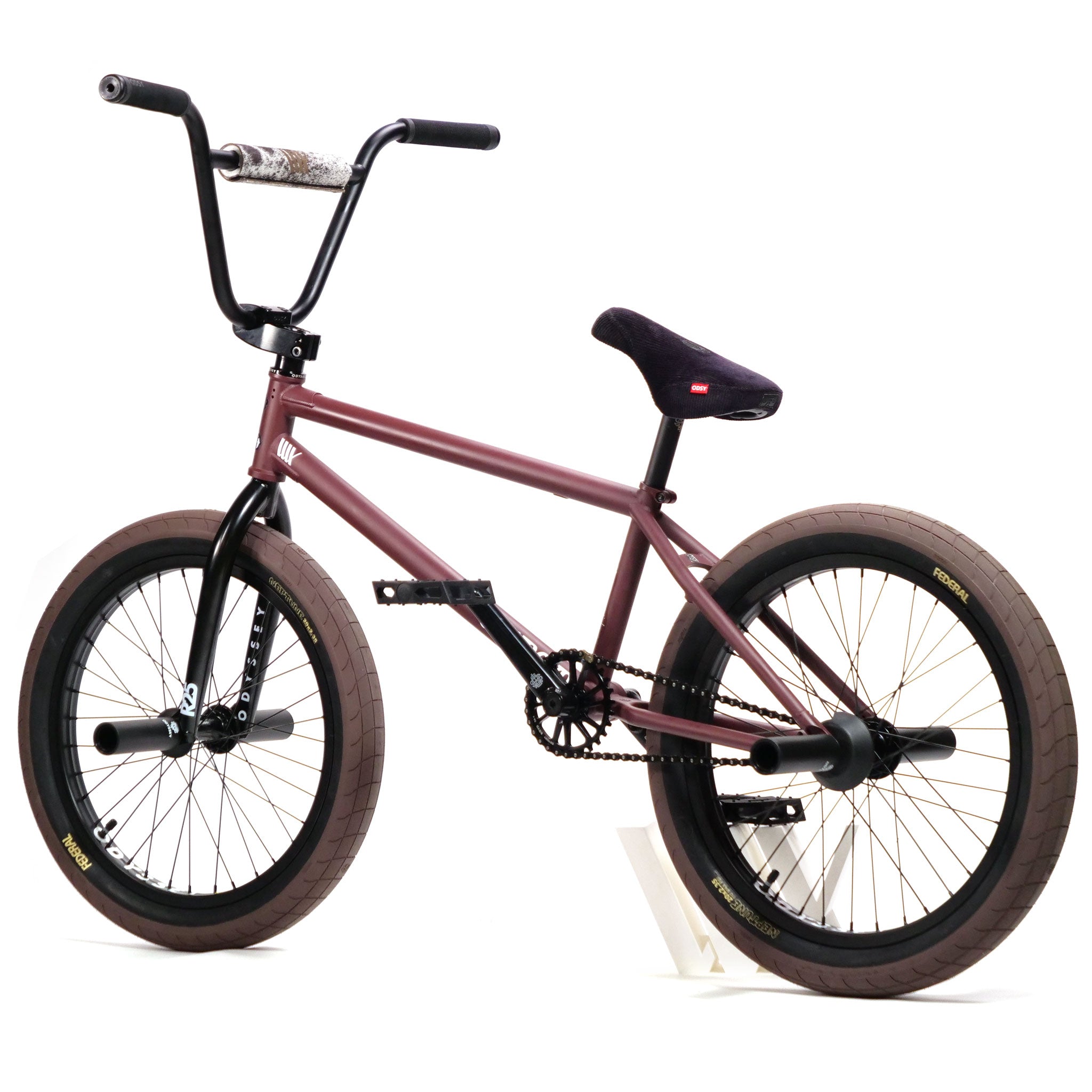 The Federal CowBoyd Custom 20 Inch Bike, with its maroon frame, rugged brown tires from Federal, and sleek black seat, stands proudly on its white stand, ready for an adventurous ride.