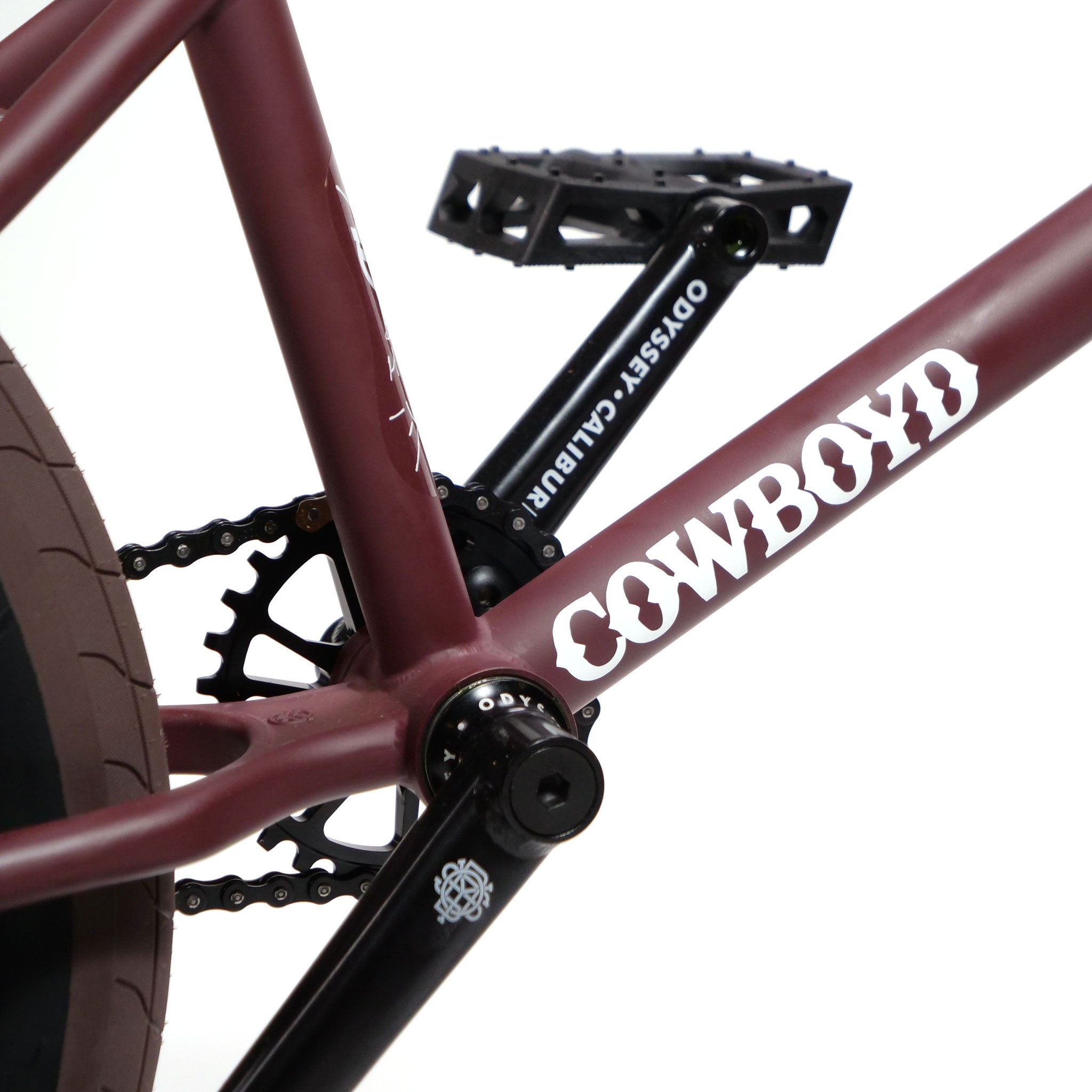 Close-up of the Federal CowBoyd Custom 20 Inch Bike's crank and chain area. The maroon frame features "COWBOY" in bold white text. Enhancing its sleekness, the bike showcases black pedals and Federal Odyssey Calibur crank arms, while a tan tire completes its stylish appearance.