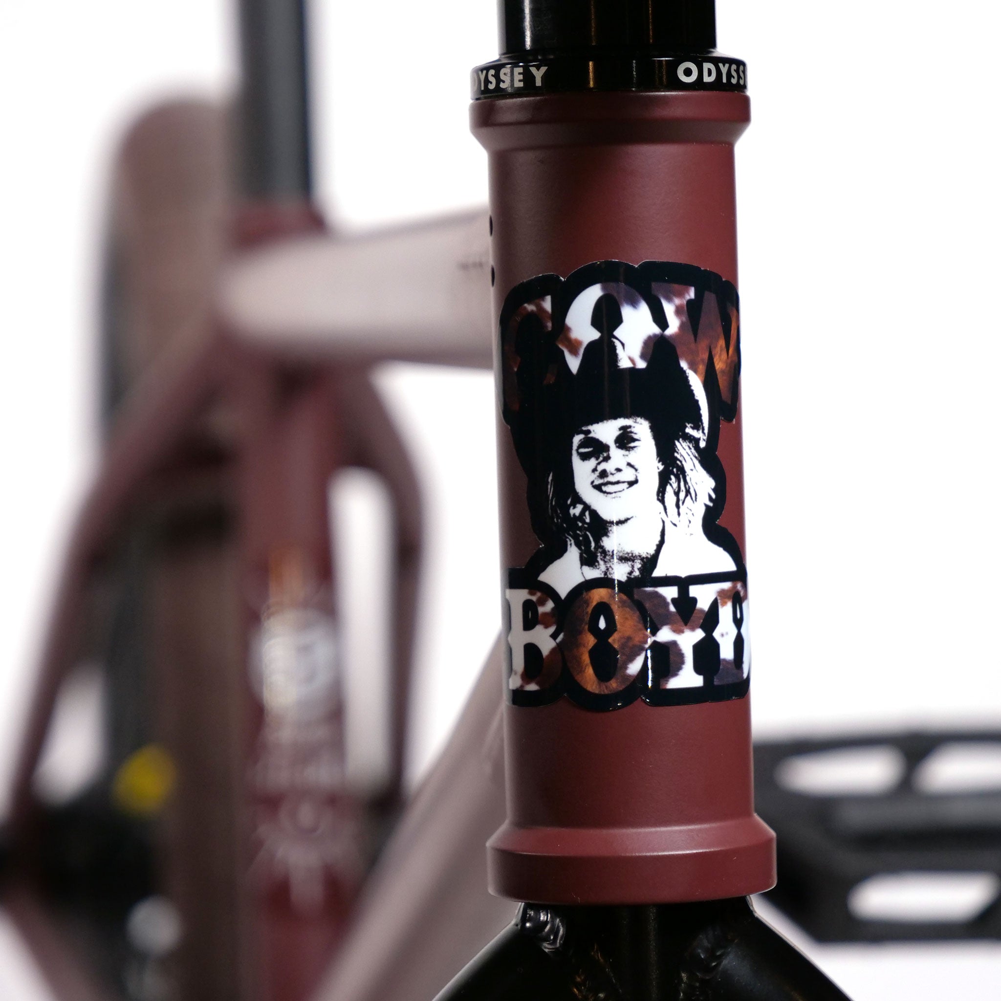 A close-up of the head tube on the maroon Federal CowBoyd Custom 20 Inch Bike reveals a sticker featuring a black-and-white image of a person wearing a cowboy hat, labeled "CowBoyd," capturing the spirit of an odyssey.