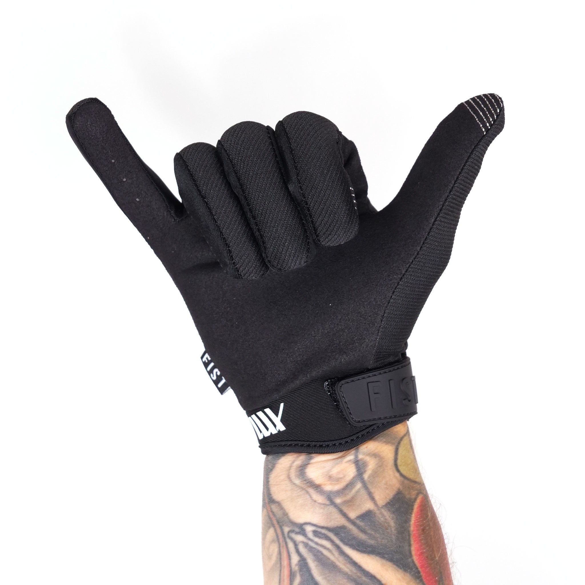 A tattooed arm wearing a black LUXBMX X FIST V2 Glove makes a shaka hand gesture against a plain white background, highlighting the seamless blend of style and function.