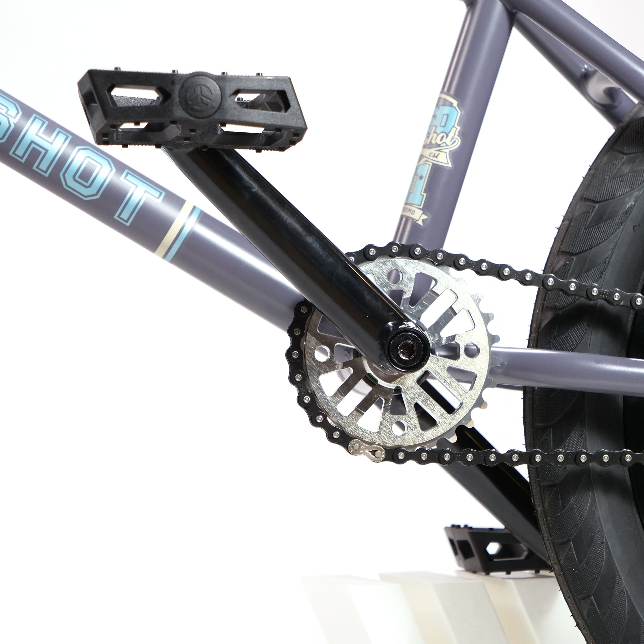 A close-up of the pedal, chainring, and chain on the BSD Long Shot Custom BMX Bike. The frame is purple with decals, and a partial view of the wheel is visible on the right.